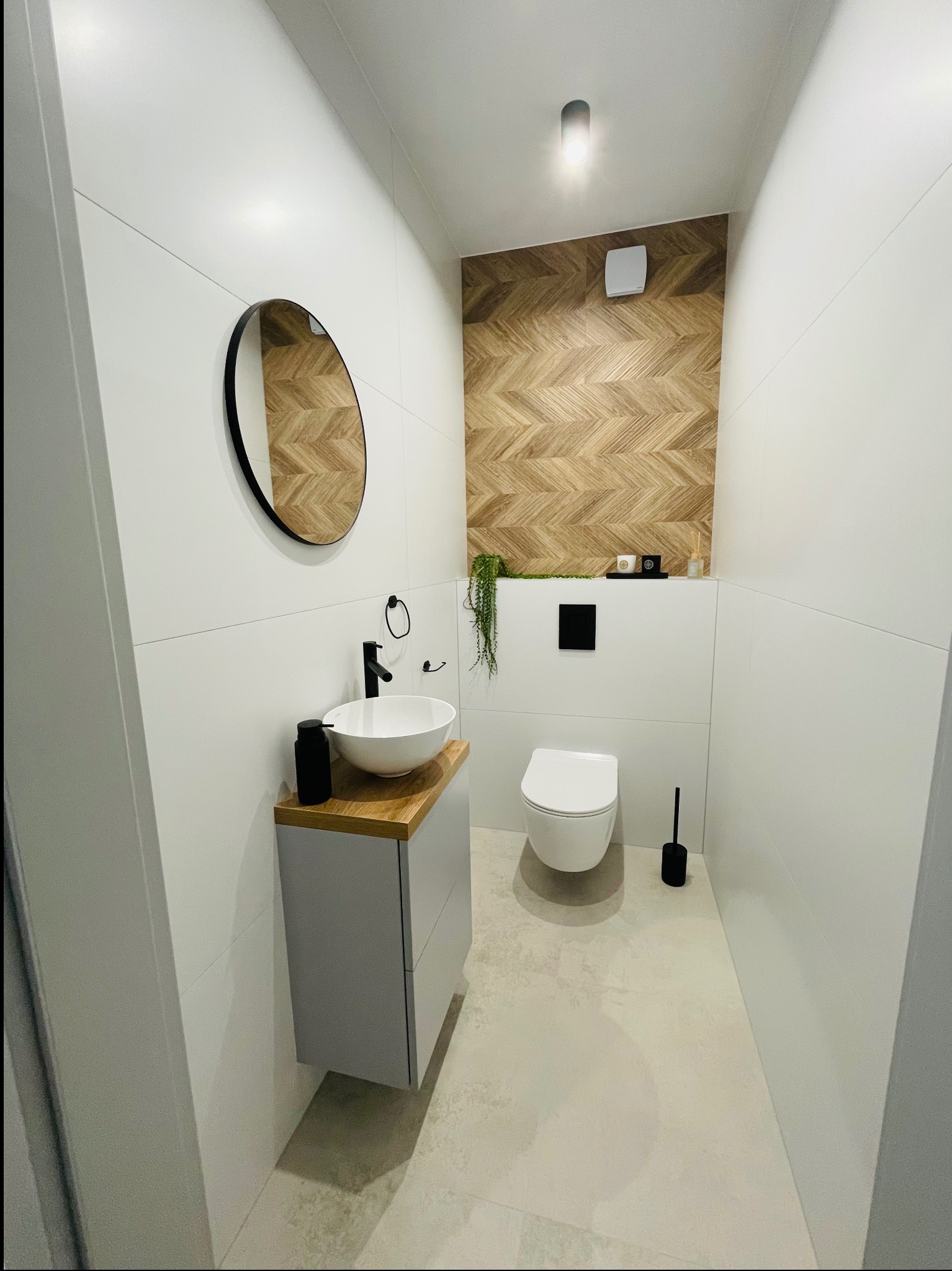 Nowoczesna toaleta z białymi płytkami, drewnianą ścianą w jodełkę, okrągłym lustrem, umywalką nablatową i szarą szafką pod umywalką.