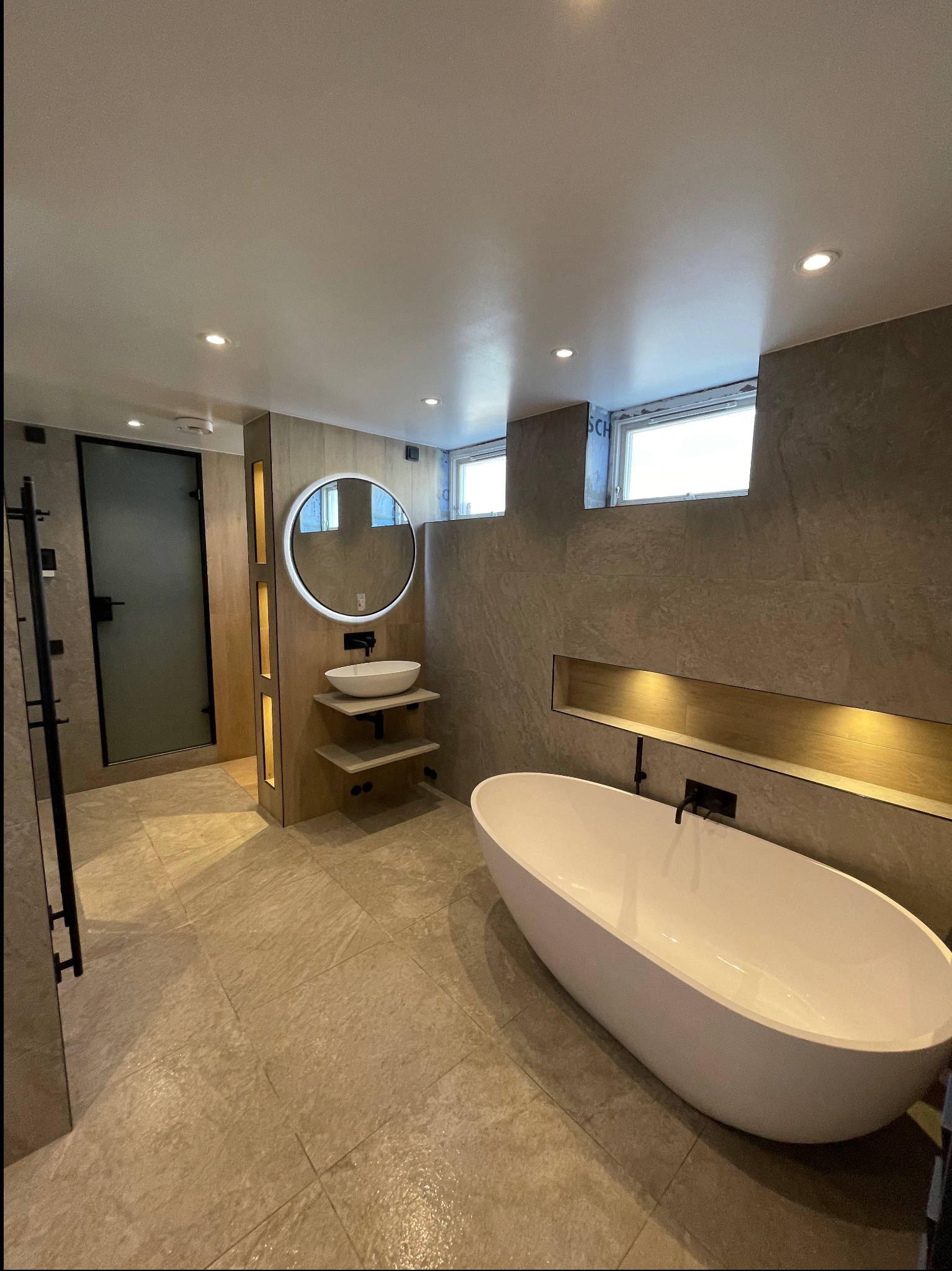 Nowoczesna łazienka z wolnostojącą wanną, okrągłym lustrem z podświetleniem LED, umywalką nablatową na drewnianych półkach i szklaną kabiną prysznicową z czarnymi elementami.