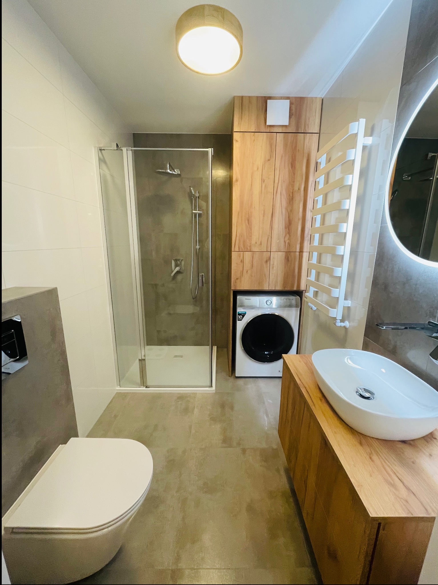 Nowoczesna łazienka z kabiną prysznicową, umywalką nablatową na drewnianej szafce, pralką w zabudowie i grzejnikiem drabinkowym.