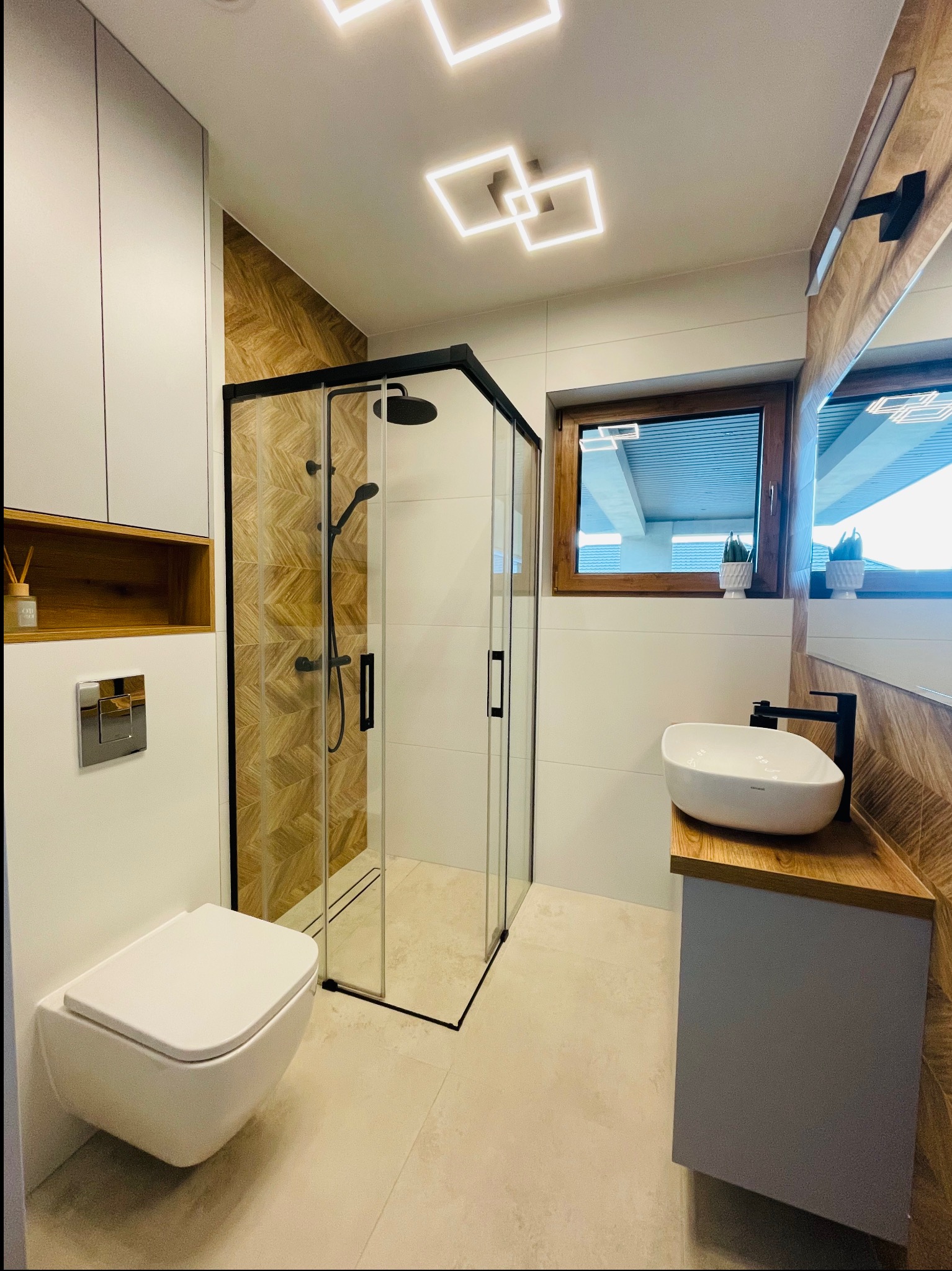 Nowoczesna łazienka z kabiną prysznicową w czarnej ramie, podwieszaną toaletą, umywalką nablatową na drewnianym blacie i geometrycznym oświetleniem sufitowym.