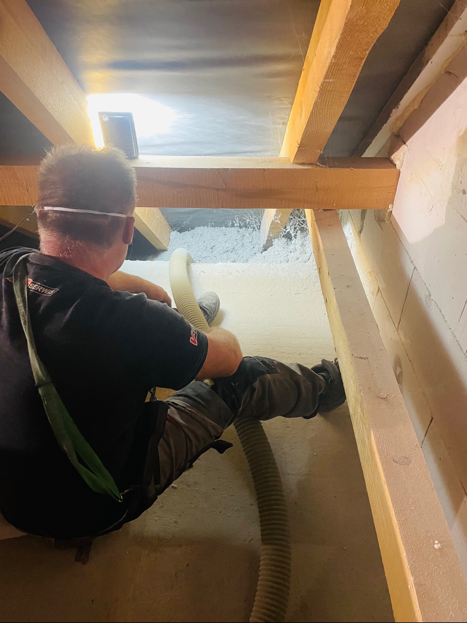 Pracownik w czarnej koszulce z logo, w okularach ochronnych, aplikuje izolację z włókien w poddaszu, używając węża i specjalistycznego sprzętu. Widoczne drewniane belki konstrukcyjne i częściowo...