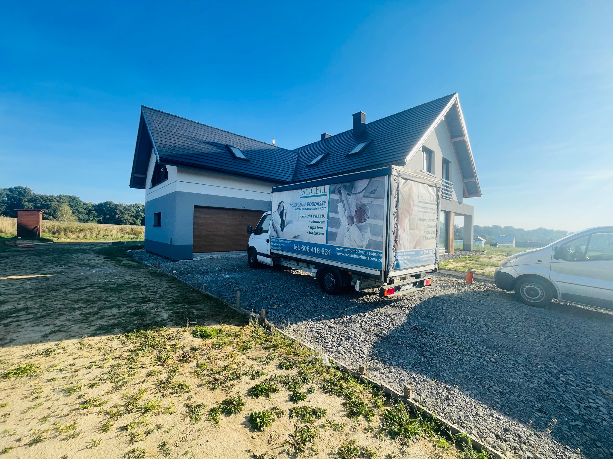 Nowoczesny dom jednorodzinny z szarą elewacją i ciemnym dachem, przed którym stoi biała furgonetka z reklamą firmy specjalizującej się w ocieplaniu poddaszy metodą wdmuchiwania celulozy, widoczny...