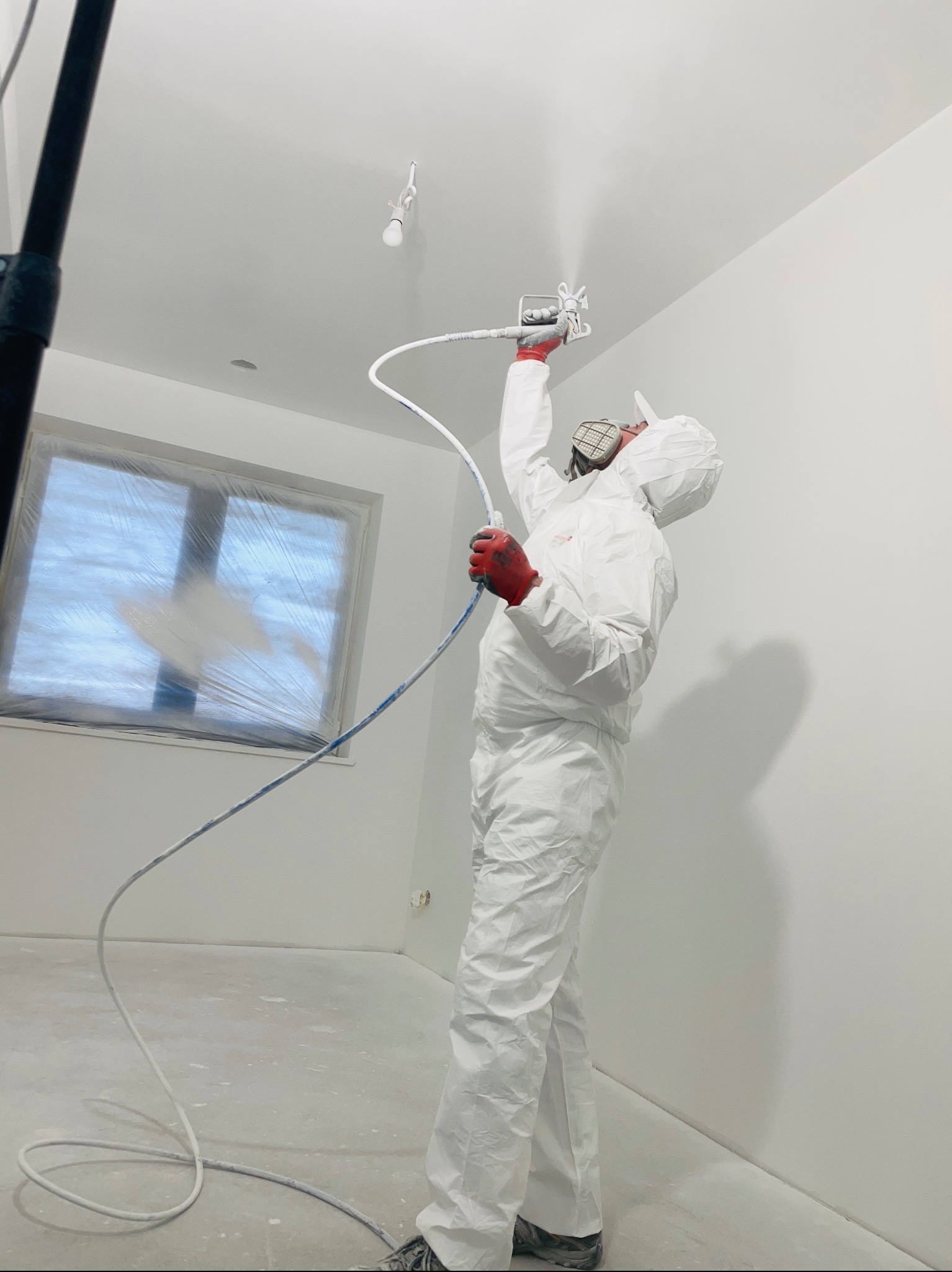 Pracownik w kombinezonie ochronnym i czerwonych rękawicach maluje sufit agregatem natryskowym; okno zabezpieczone folią malarską.