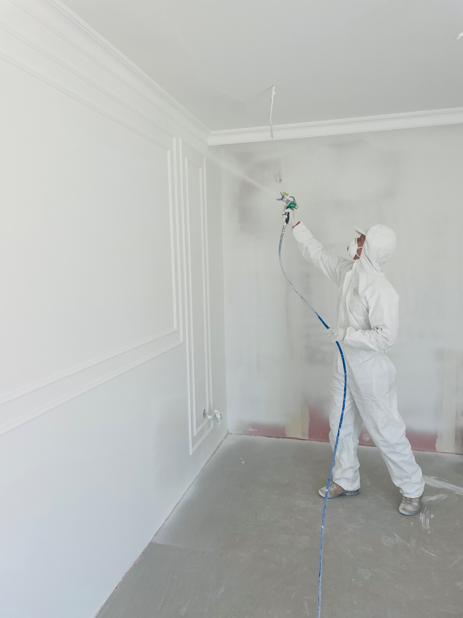 Malowanie natryskowe ścian z dekoracyjnymi listwami w Iwierzycach. Pracownik w kombinezonie nanosi białą farbę na ścianę za pomocą pistoletu.