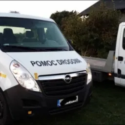 Dwa białe busy pomocy drogowej z napisem 'Pomoc Drogowa' i numerem telefonu, zaparkowane na trawniku przed domem.