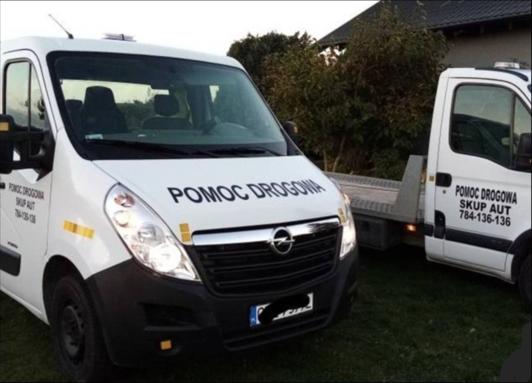 Dwa białe busy pomocy drogowej z napisem 'Pomoc Drogowa' i numerem telefonu, zaparkowane na trawniku przed domem.