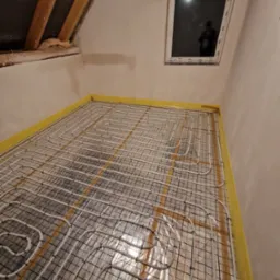 Instalacja ogrzewania podłogowego w nowym domu, widoczne rury grzewcze ułożone na siatce, żółta taśma izolacyjna przy ścianach, okno dachowe w tle, surowe ściany.