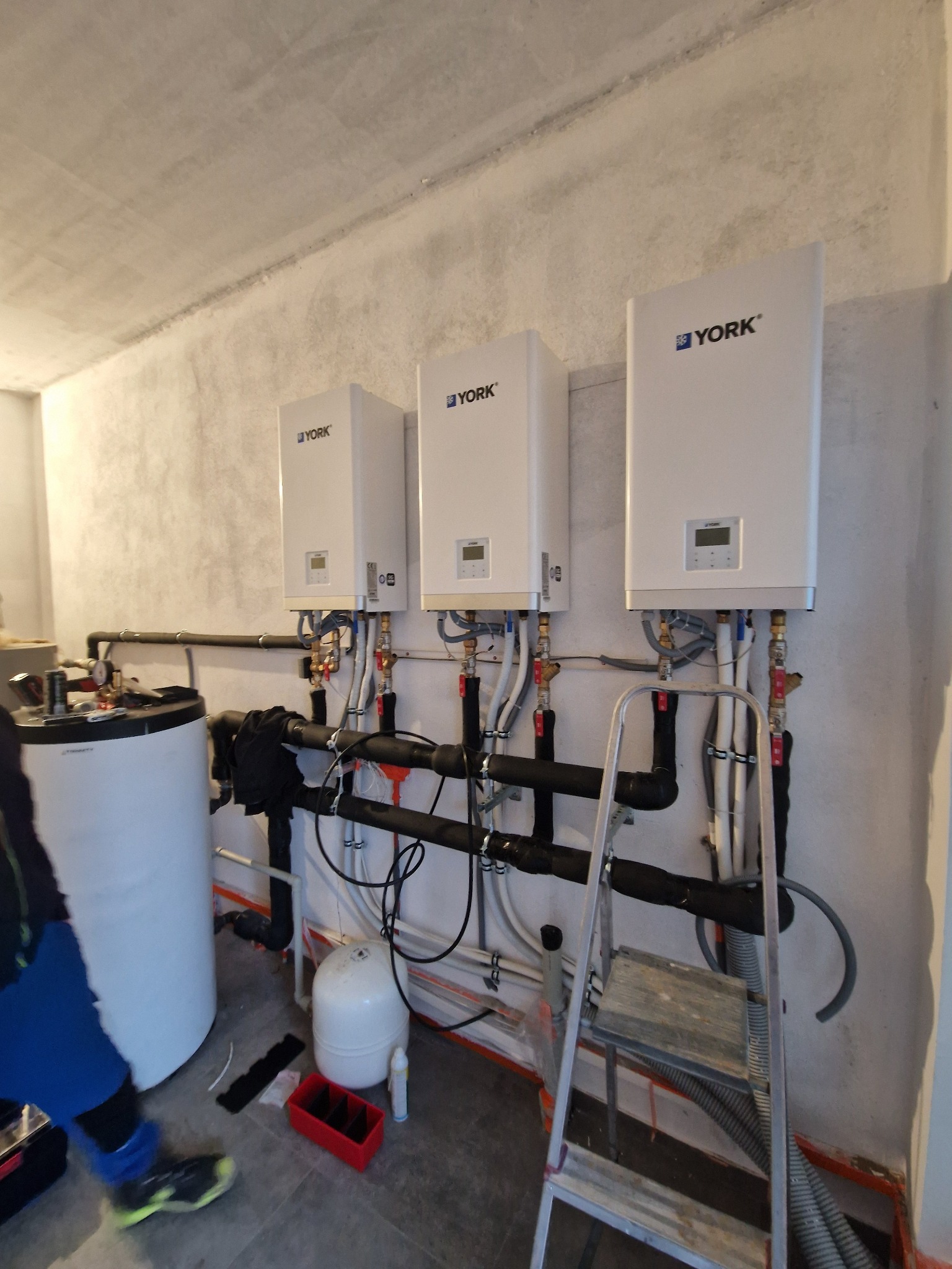 Trzy wiszące, białe kotły grzewcze marki York, podłączone rurami instalacji grzewczej, drabina oparta o ścianę, instalacja w surowym pomieszczeniu.
