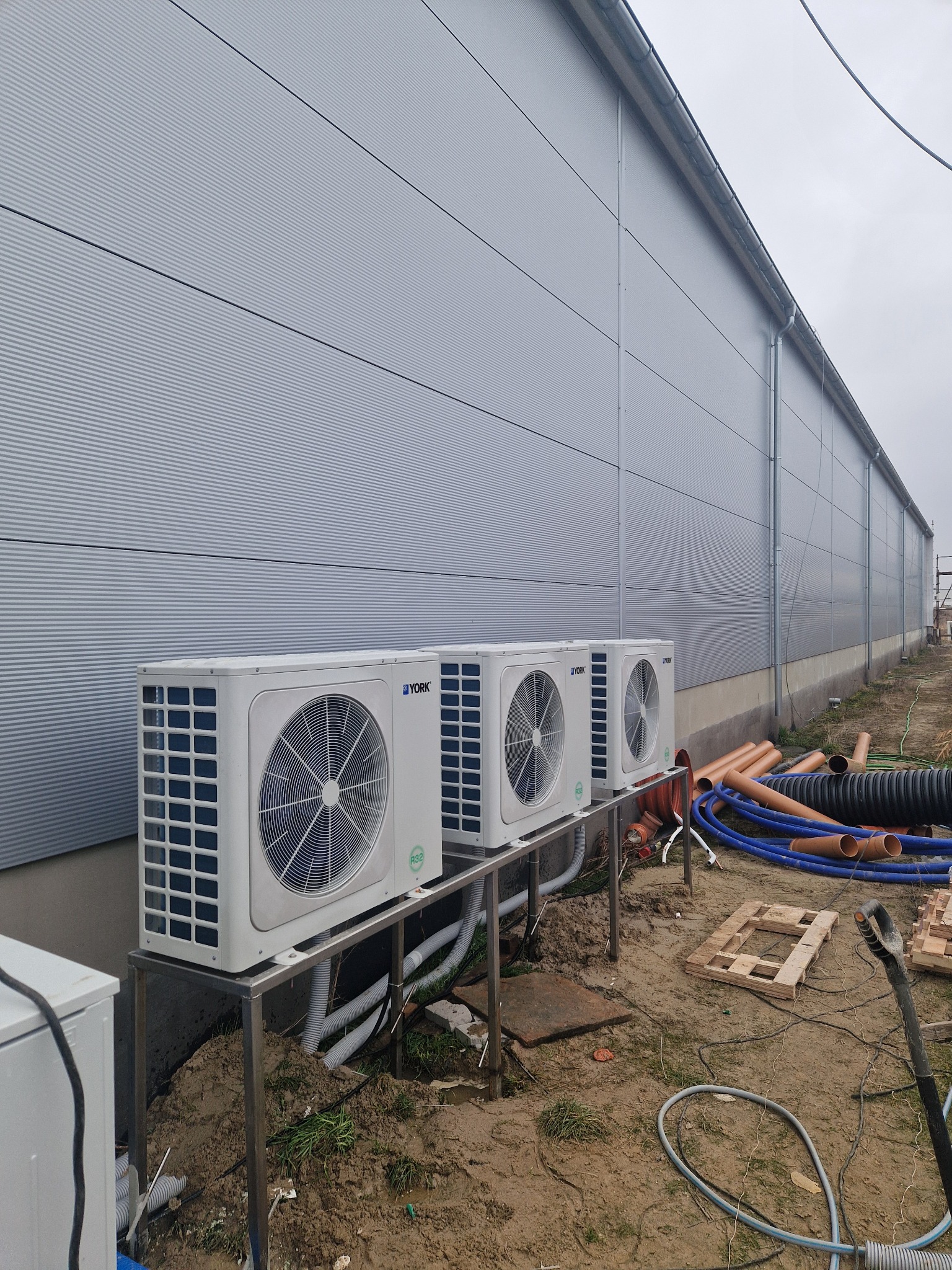 Trzy jednostki zewnętrzne klimatyzacji marki York, zamontowane na metalowym stelażu przy szarej hali magazynowej z widocznymi rurami i elementami instalacji na ziemi.