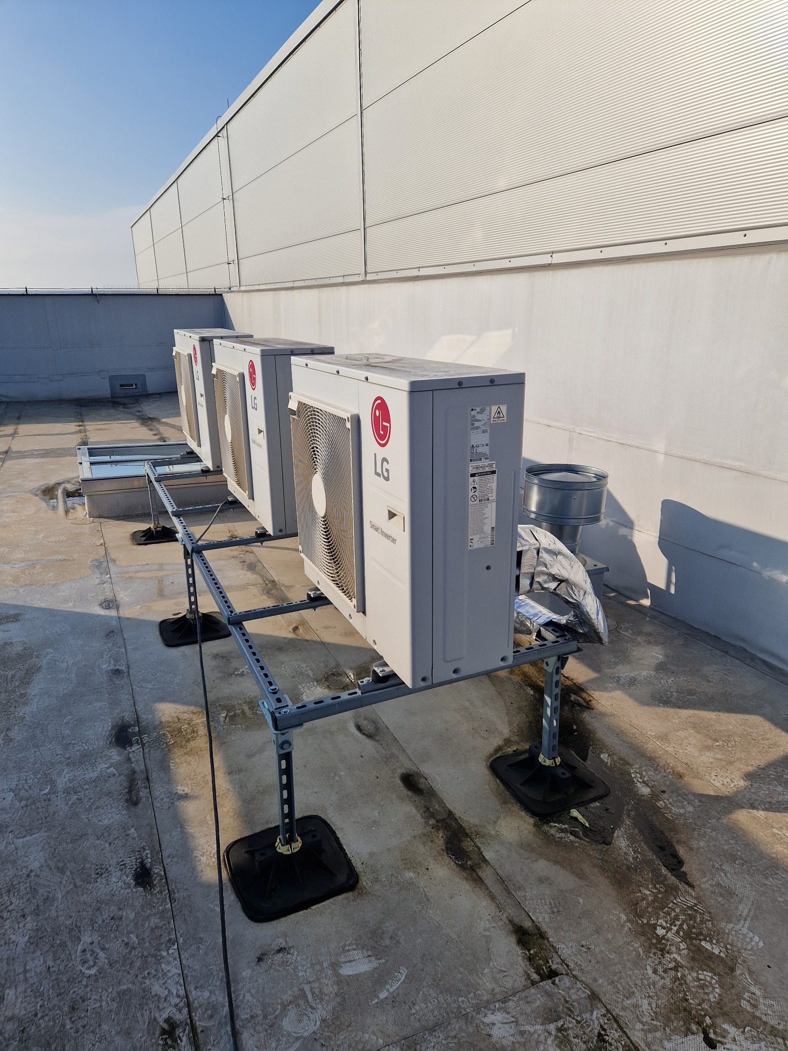 Trzy jednostki zewnętrzne klimatyzacji LG na dachu budynku w Obra, zamontowane na metalowych konstrukcjach wsporczych z czarnymi podstawami, z widocznym fragmentem instalacji wentylacyjnej i ścianą...