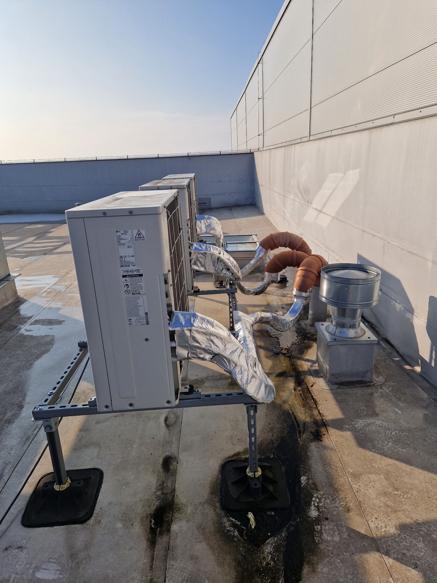 Rząd klimatyzatorów na dachu budynku przemysłowego, z widocznymi przewodami w srebrnej izolacji i elementami wentylacyjnymi, na tle jasnego nieba.