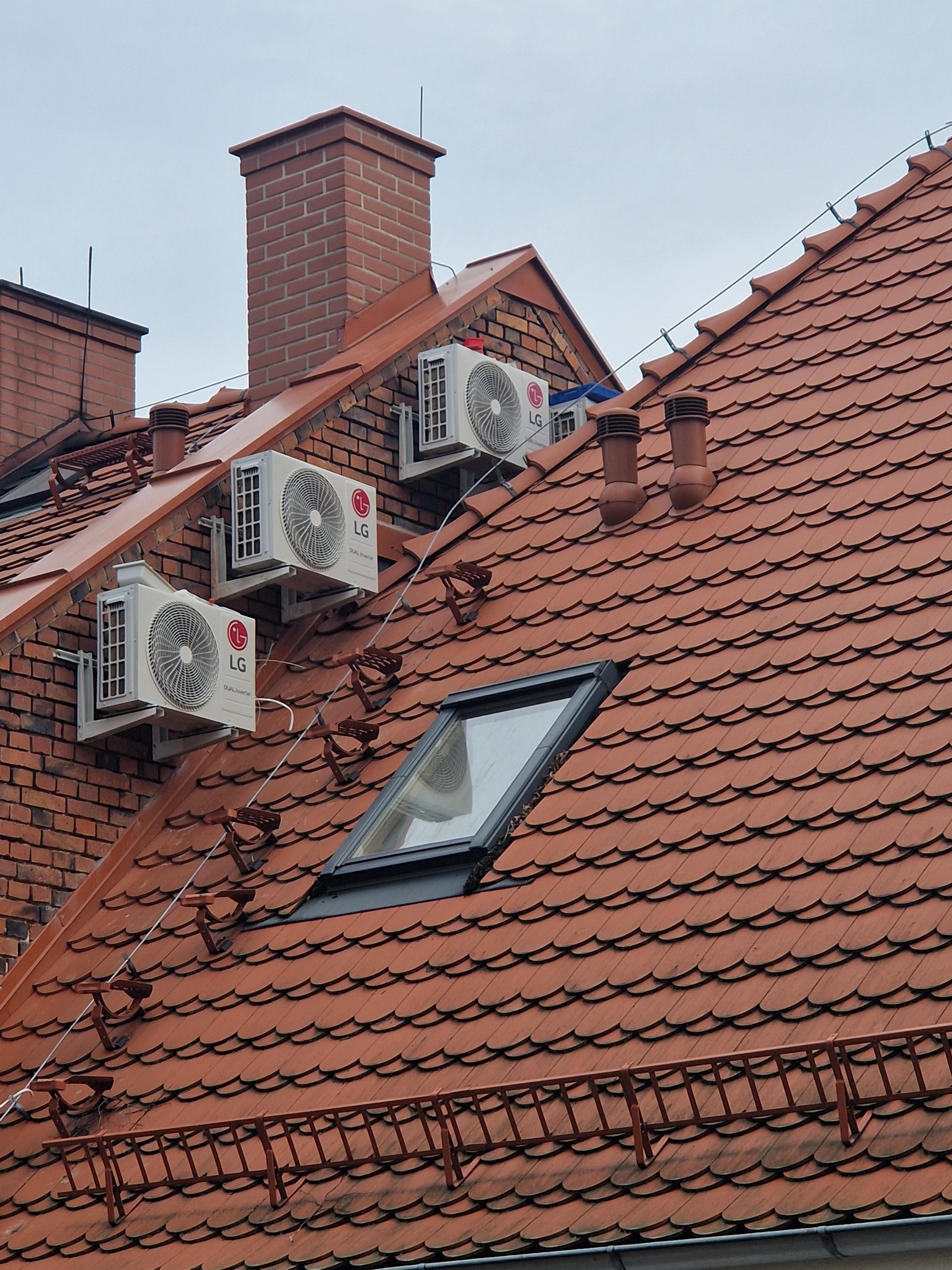 Trzy klimatyzatory LG zamontowane na dachu z czerwonej dachówki w pobliżu okna dachowego i ceglanego komina. Widoczne uchwyty do mocowania klimatyzatorów i zabezpieczenia na dachu.