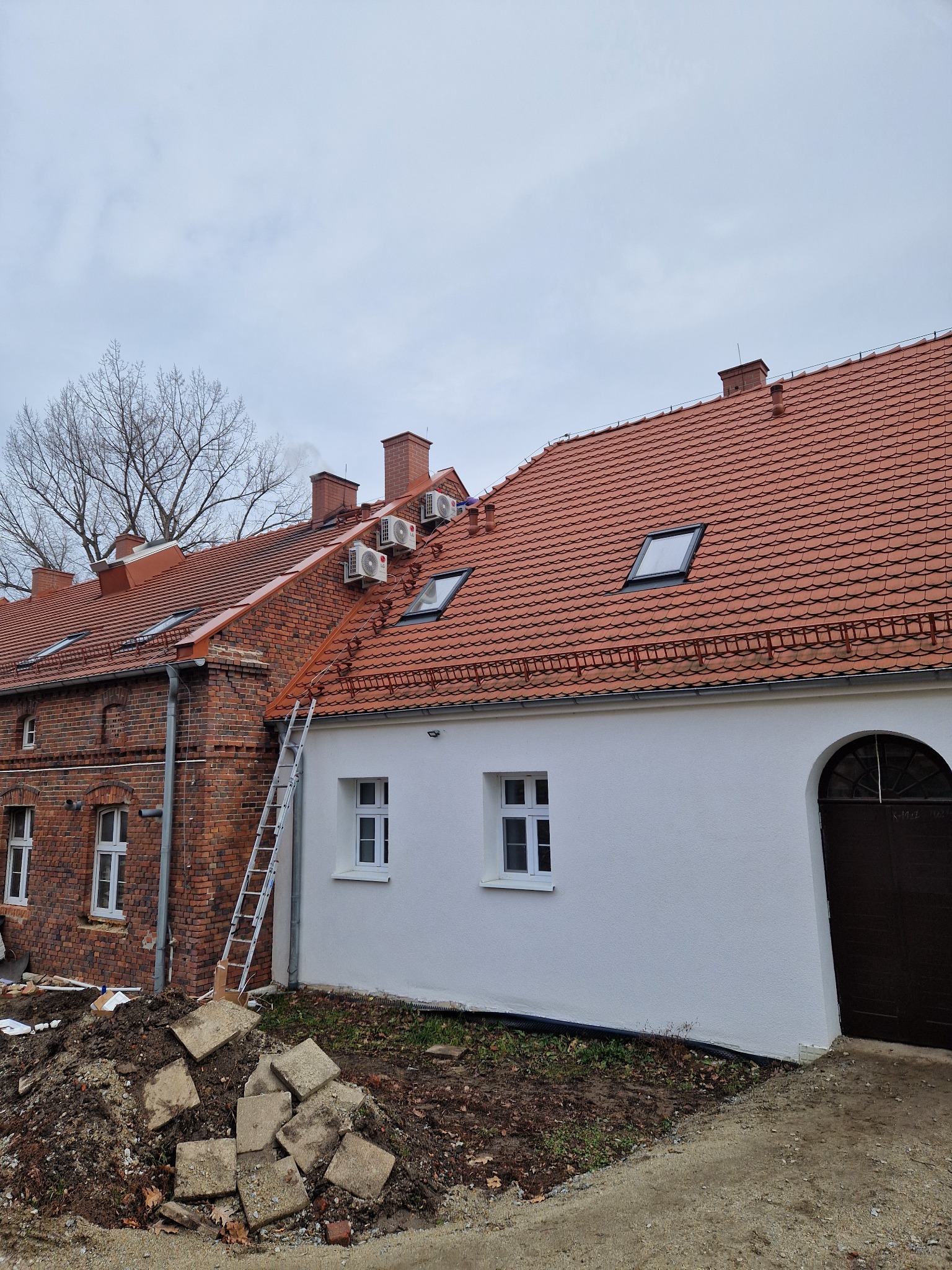Montaż klimatyzatorów LG na dachu budynku z czerwoną dachówką, widoczna aluminiowa drabina oparta o ścianę. Fragment budynku z cegły i fragment z białą elewacją.