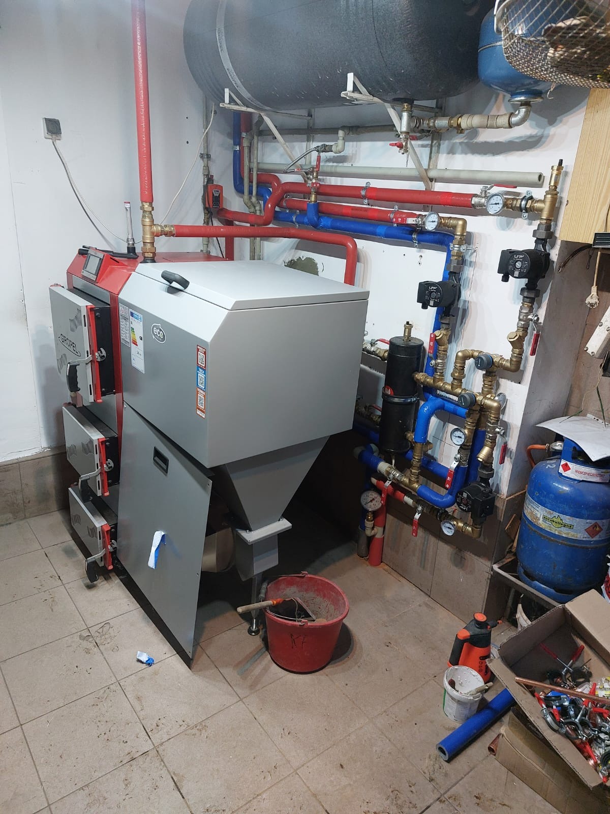 Nowoczesny piec na paliwo stałe z zasobnikiem, z widocznymi rurami instalacji grzewczej w kolorach czerwonym i niebieskim, oraz osprzętem, zamontowany w kotłowni z ceramiczną podłogą.