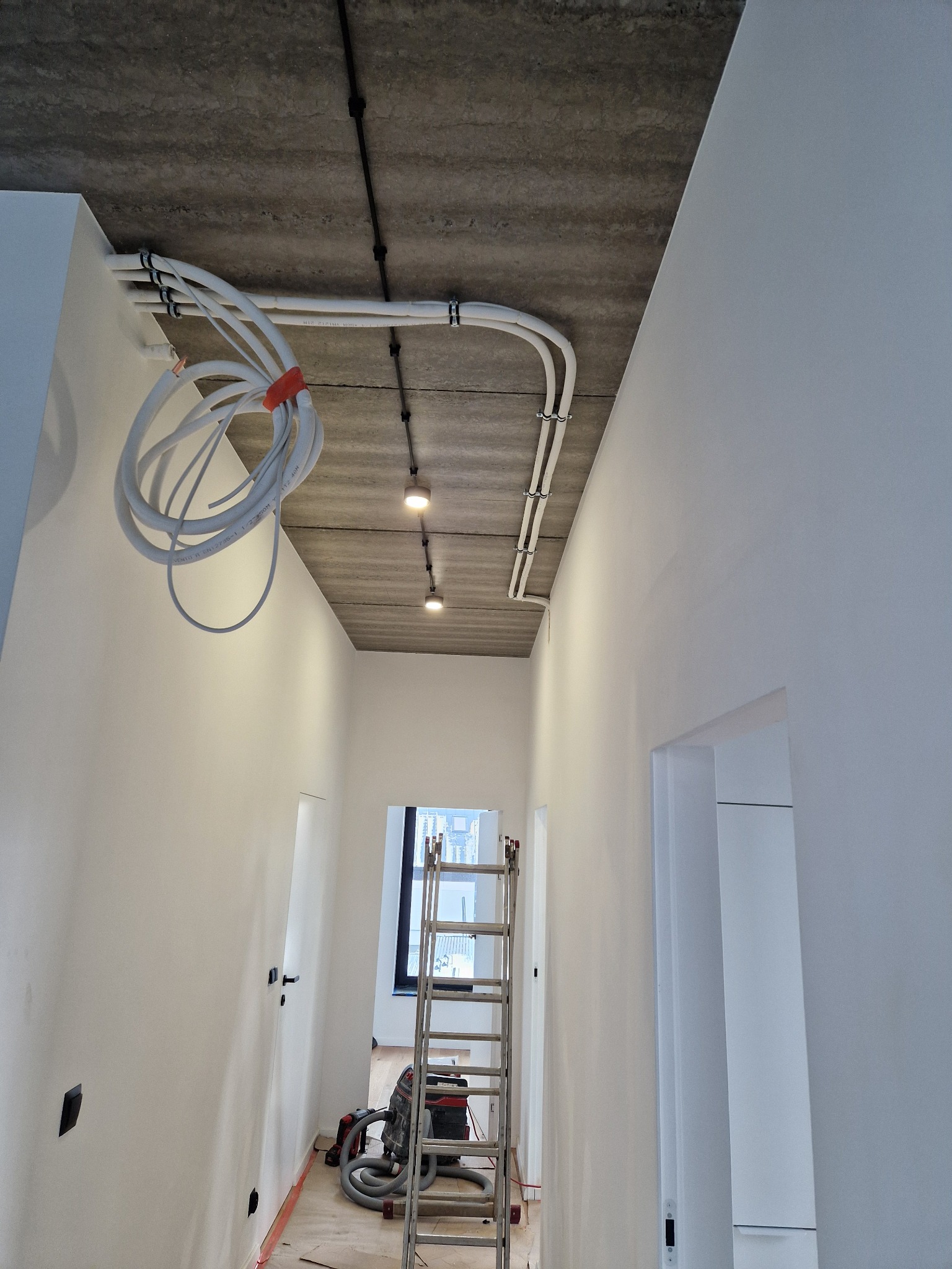 Wnętrze w trakcie remontu: białe ściany, surowy betonowy sufit z zamontowanymi lampami punktowymi i wiązką białych kabli elektrycznych z pomarańczową opaską, drabina oparta o ścianę, widoczny...