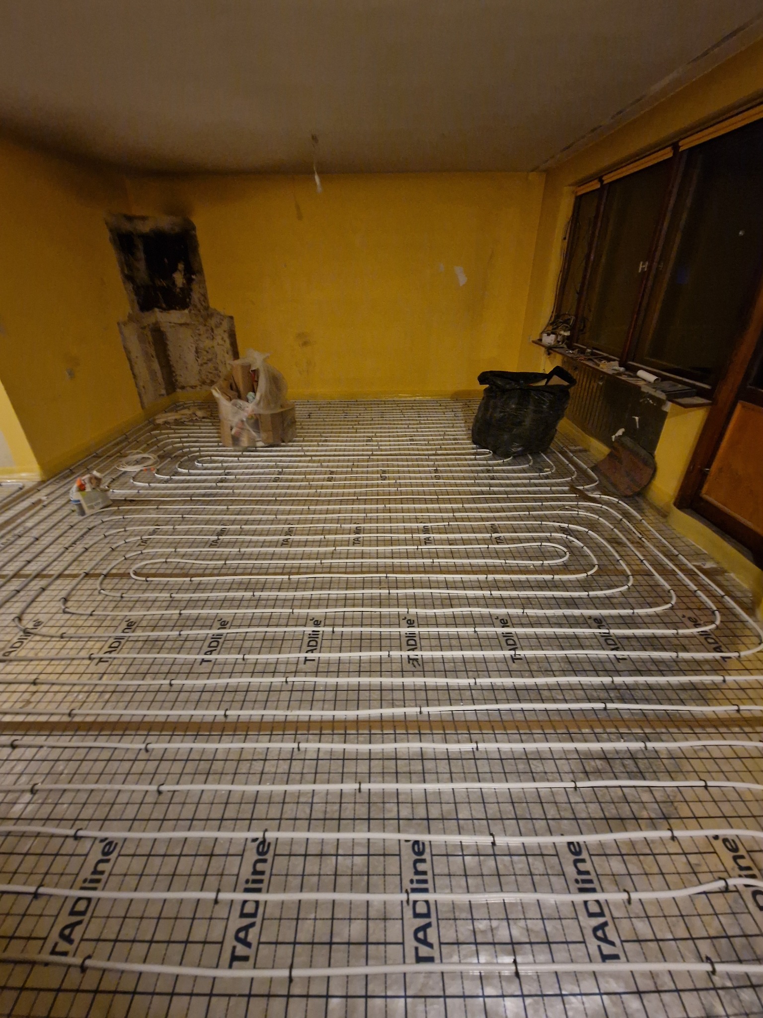 Rozłożona instalacja ogrzewania podłogowego TADline na siatce, widoczne białe rury układane w pętle na żółtej podłodze w trakcie prac, w tle ściana z otworem po kominku i okno.