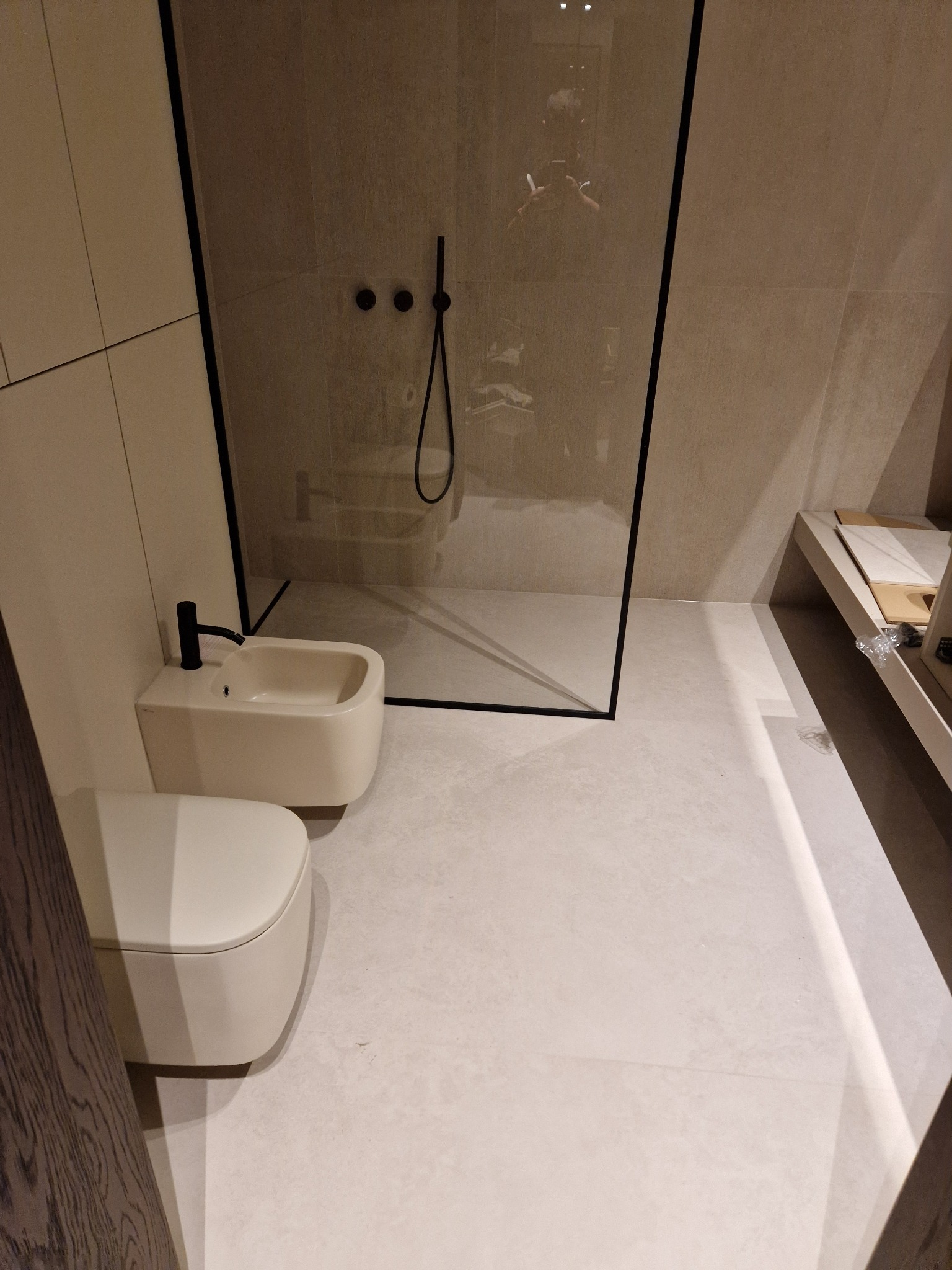 Nowoczesna łazienka z beżowymi płytkami, kabiną prysznicową typu walk-in z czarnymi profilami, wiszącą toaletą i bidetem z czarną armaturą.