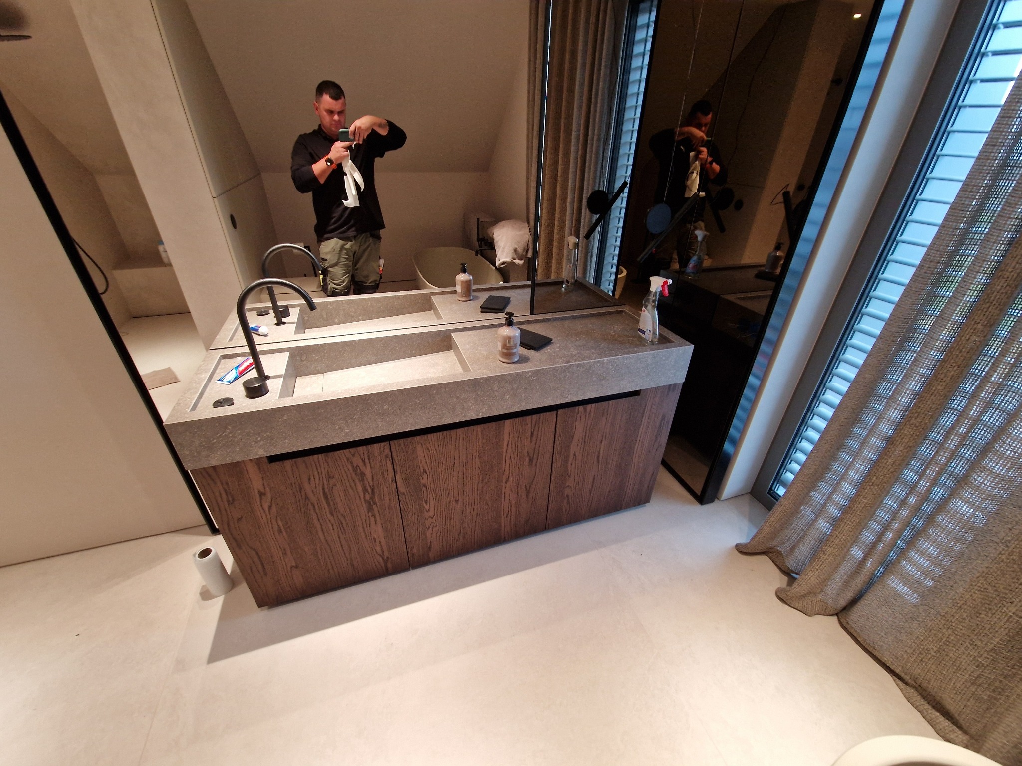 Mężczyzna w czarnej koszulce czyści armaturę umywalkową w nowoczesnej łazience z podwójną umywalką i drewnianą szafką podblatową.