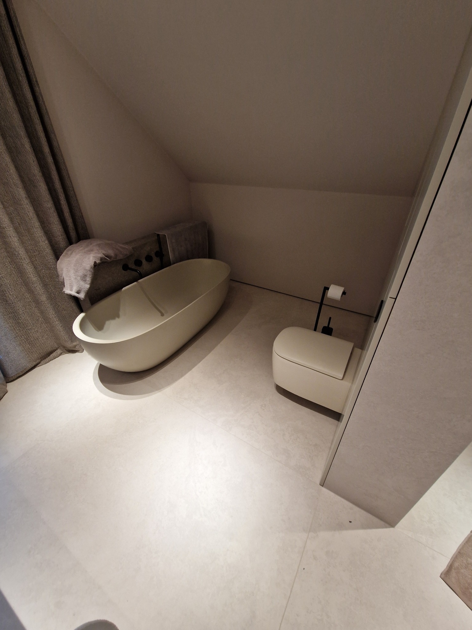 Minimalistyczna łazienka na poddaszu z owalną wanną wolnostojącą, podtynkową armaturą, wiszącą toaletą i neutralną kolorystyką.