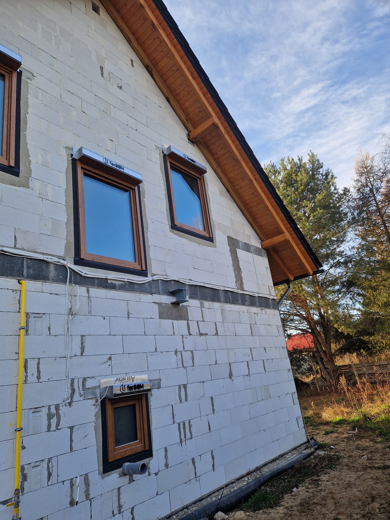 Niedokończona elewacja domu z pustaków, widoczne okna z drewnianą ramą i szare rolety, żółta rura gazowa biegnąca wzdłuż ściany, instalacja elektryczna na zewnątrz.