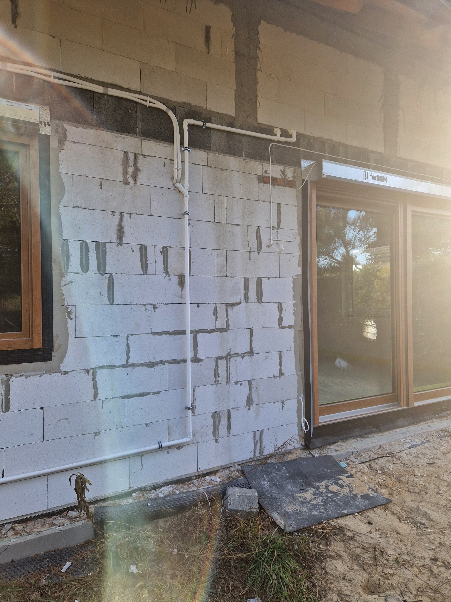 Białe rury instalacyjne prowadzone po zewnętrznej ścianie budynku z betonu komórkowego, widoczne okna z drewnianymi ramami, teren wokół budynku w trakcie prac wykończeniowych.