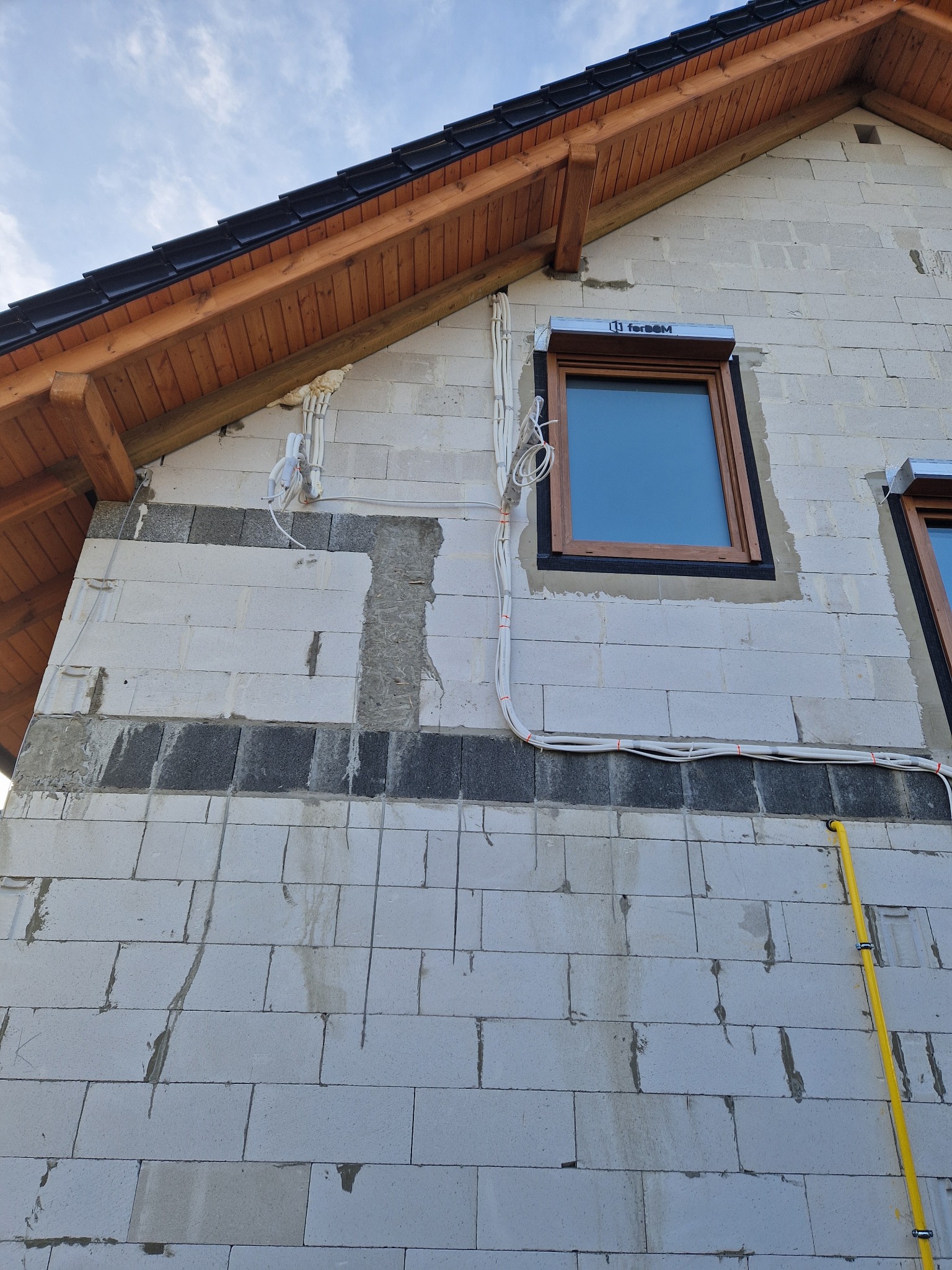 Ściana zewnętrzna budynku z widocznymi przewodami instalacji elektrycznej i rurą gazową, okna z roletami zewnętrznymi, stan surowy.