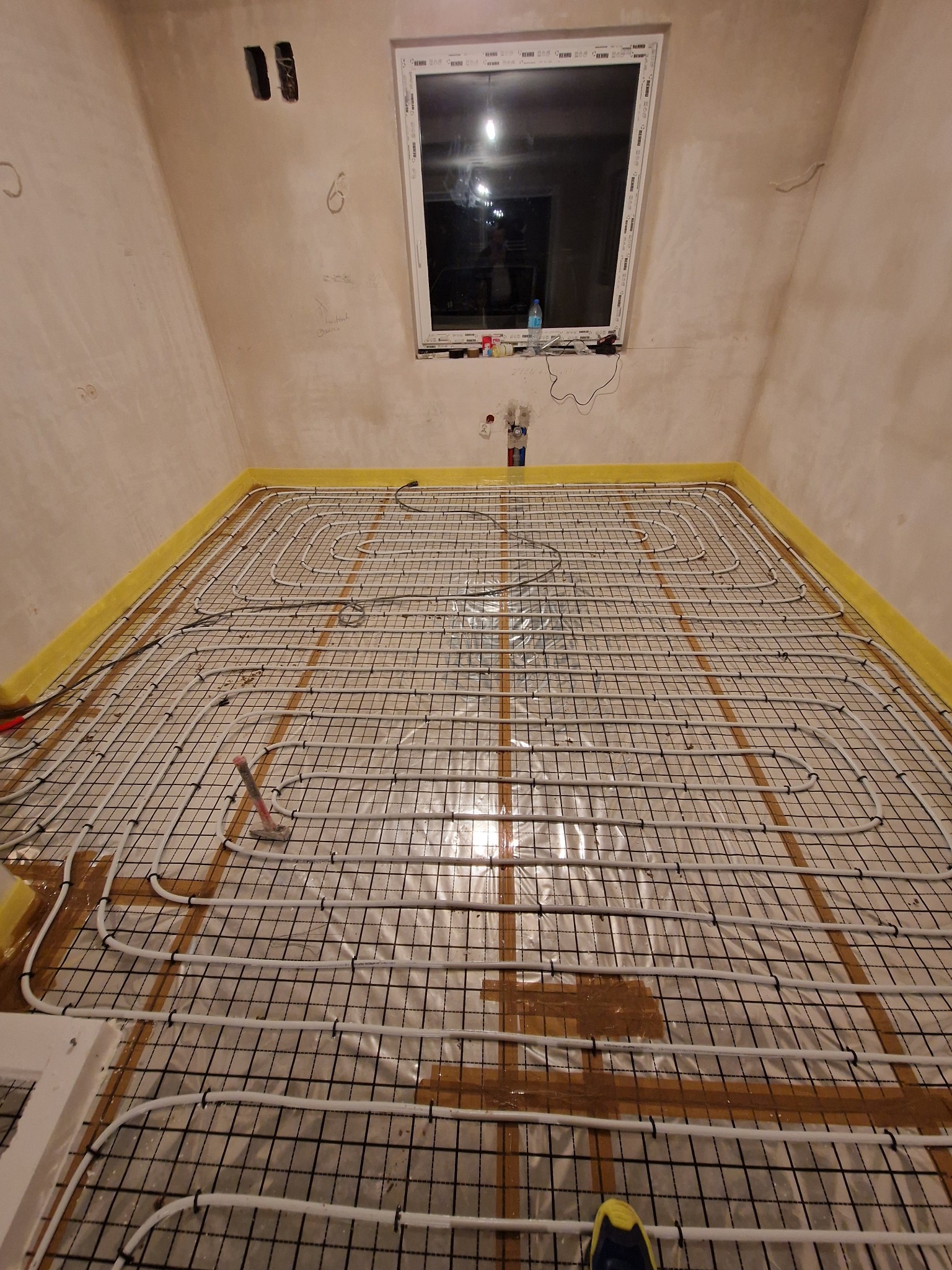 Montaż ogrzewania podłogowego: białe rury grzewcze ułożone na czarnej siatce montażowej, żółta taśma brzegowa przy ścianach, surowy stan ścian i okno w tle.