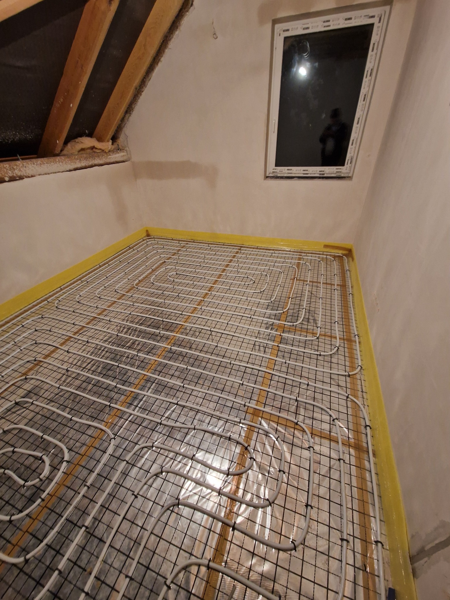 Instalacja ogrzewania podłogowego w nowym domu, widoczne rury grzewcze ułożone na siatce, żółta taśma izolacyjna przy ścianach, okno dachowe w tle, surowe ściany.