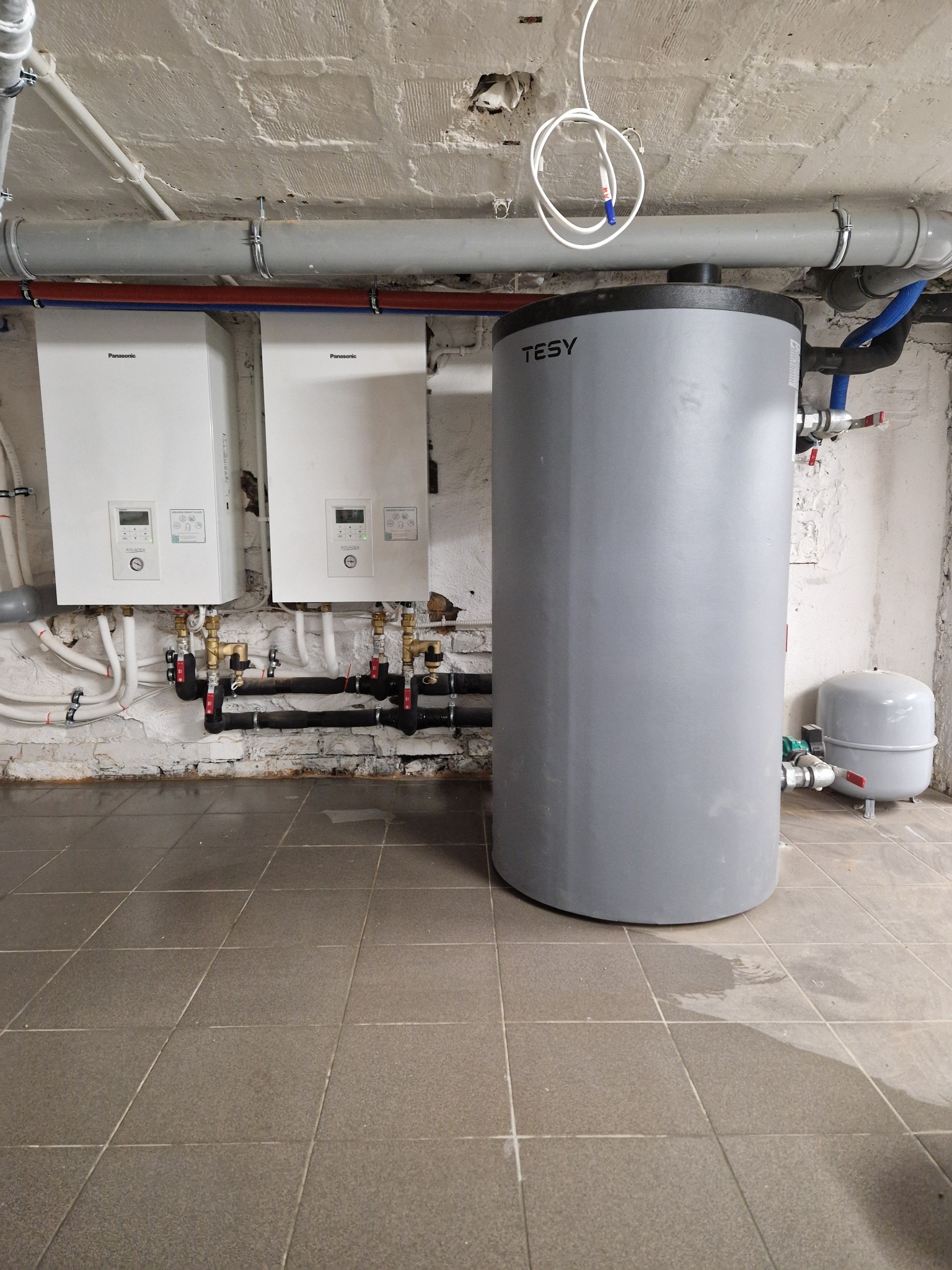 Dwa białe kotły gazowe Panasonic Aquarea, szary zbiornik akumulacyjny TESY i małe naczynie wzbiorcze w piwnicy z instalacjami, widoczne rury i armatura.