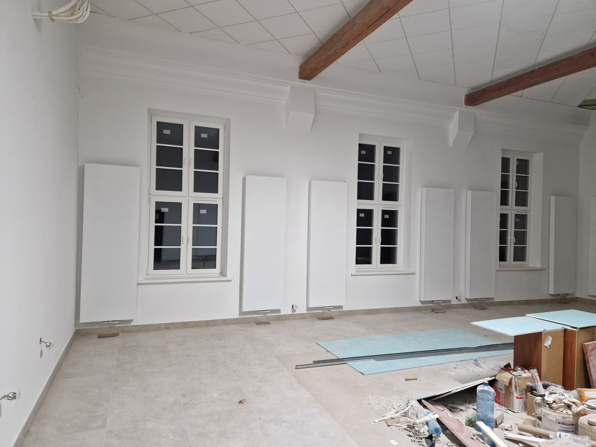 Wnętrze pomieszczenia po remoncie z nowymi, białymi grzejnikami panelowymi pod oknami, widoczne pozostałości po pracach budowlanych na podłodze i drewniane belki stropowe.