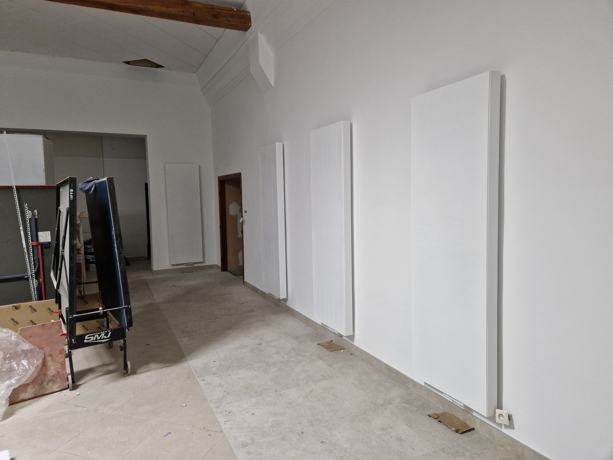 Wnętrze z zamontowanymi białymi panelami grzewczymi na ścianie, widoczna instalacja elektryczna przy podłodze oraz fragment stołu do ping-ponga w tle.