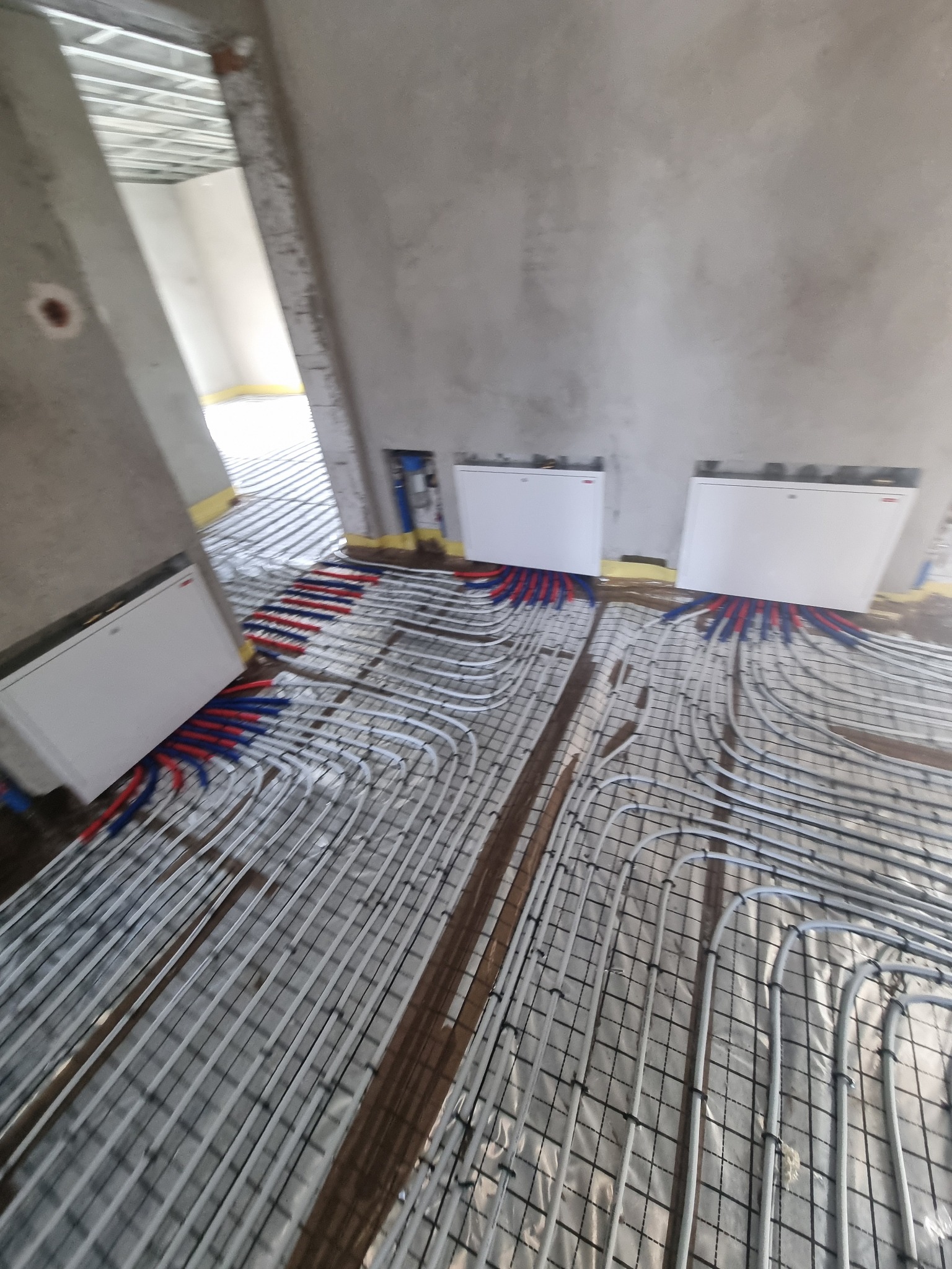 Wnętrze w trakcie instalacji ogrzewania podłogowego, widoczne białe rury grzewcze ułożone na folii izolacyjnej z siatką, podłączone do grzejników ściennych, surowy stan wykończenia ścian i sufitu.