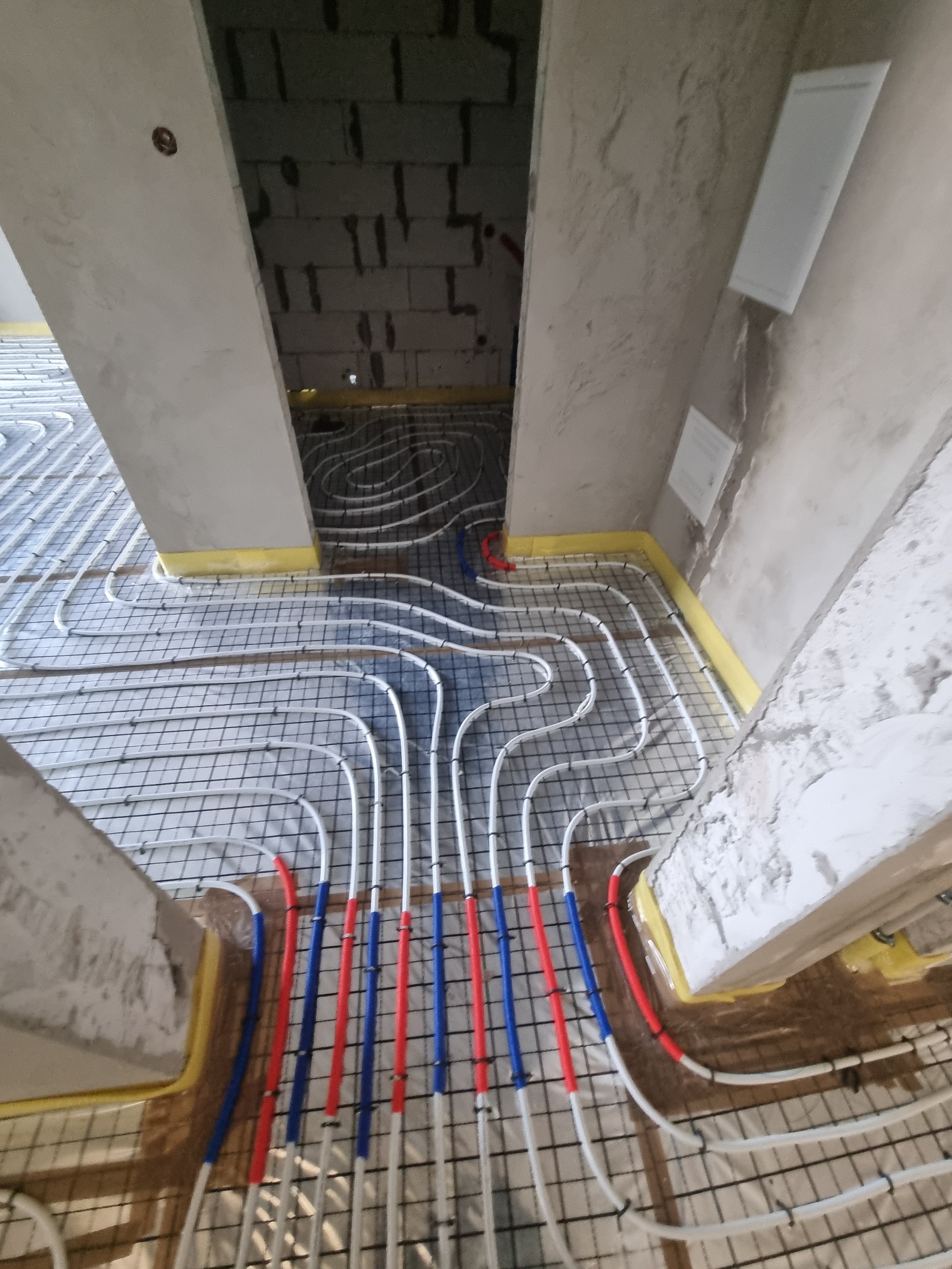 Montaż ogrzewania podłogowego: białe rurki grzewcze ułożone w pętle na metalowej siatce, częściowo pod wylewką, z widocznymi fragmentami czerwonych i niebieskich rur przy ścianach, zabezpieczonych...
