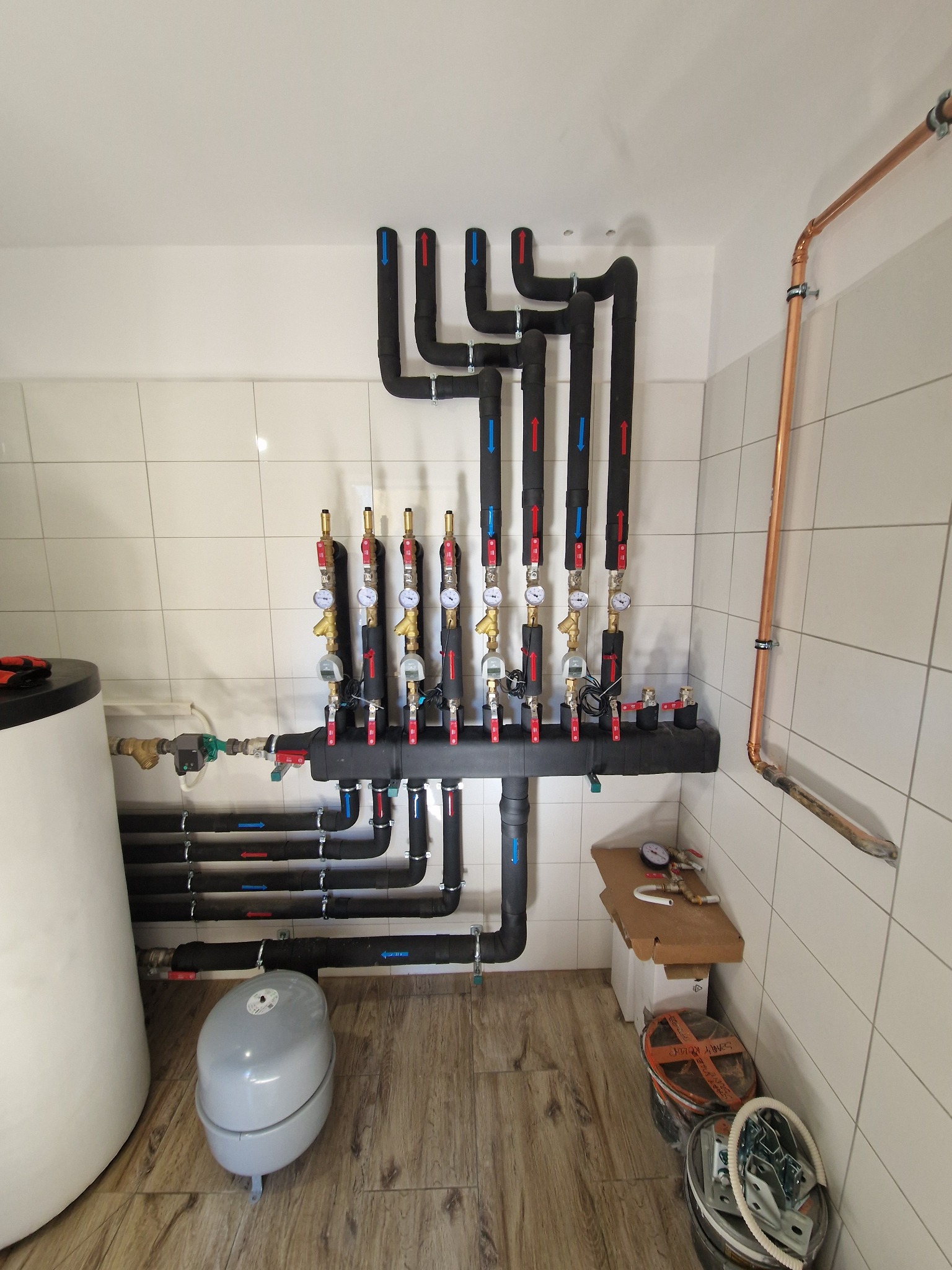Rozdzielacz instalacji grzewczej z rurami oznaczonymi strzałkami, manometry, zbiornik wyrównawczy. Instalacja w pomieszczeniu z białymi kafelkami i drewnopodobną podłogą.