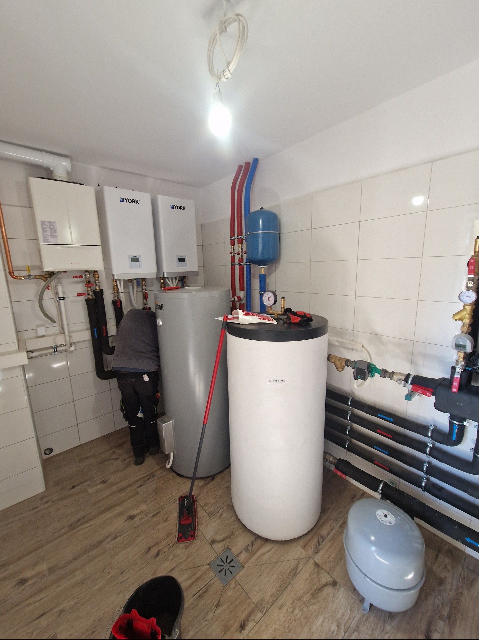 W kotłowni zamontowane bojlery i piece gazowe York, z widocznymi rurami instalacji. Jeden z instalatorów w trakcie pracy przy szarym bojlerze. Na podłodze mop i buty.