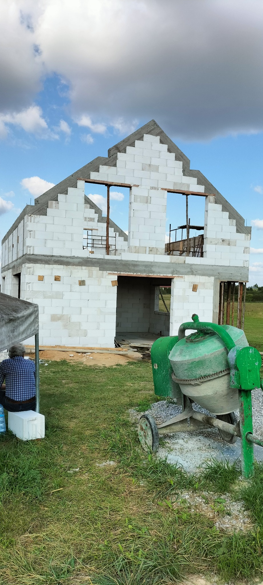 Budowa domu z białych bloczków, widoczna betoniarka i osoba odpoczywająca w cieniu. Dom w stanie surowym otwartym, z widocznymi otworami okiennymi i zarysem dachu.