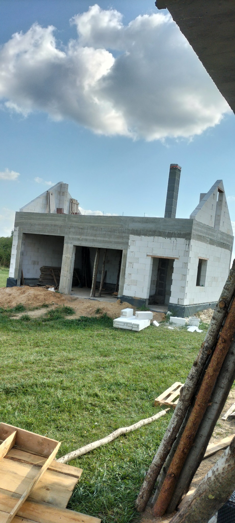 Budowa domu jednorodzinnego w stanie surowym otwartym, widoczne białe bloczki, betonowe nadproża i komin. Na pierwszym planie trawa i drewniane elementy.