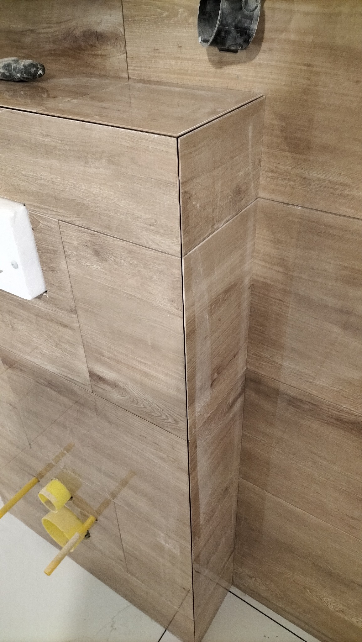 Obudowa stelaża WC wykończona płytkami imitującymi drewno, z widocznymi przyłączami hydraulicznymi. Estetyczne połączenie płytek na krawędziach.