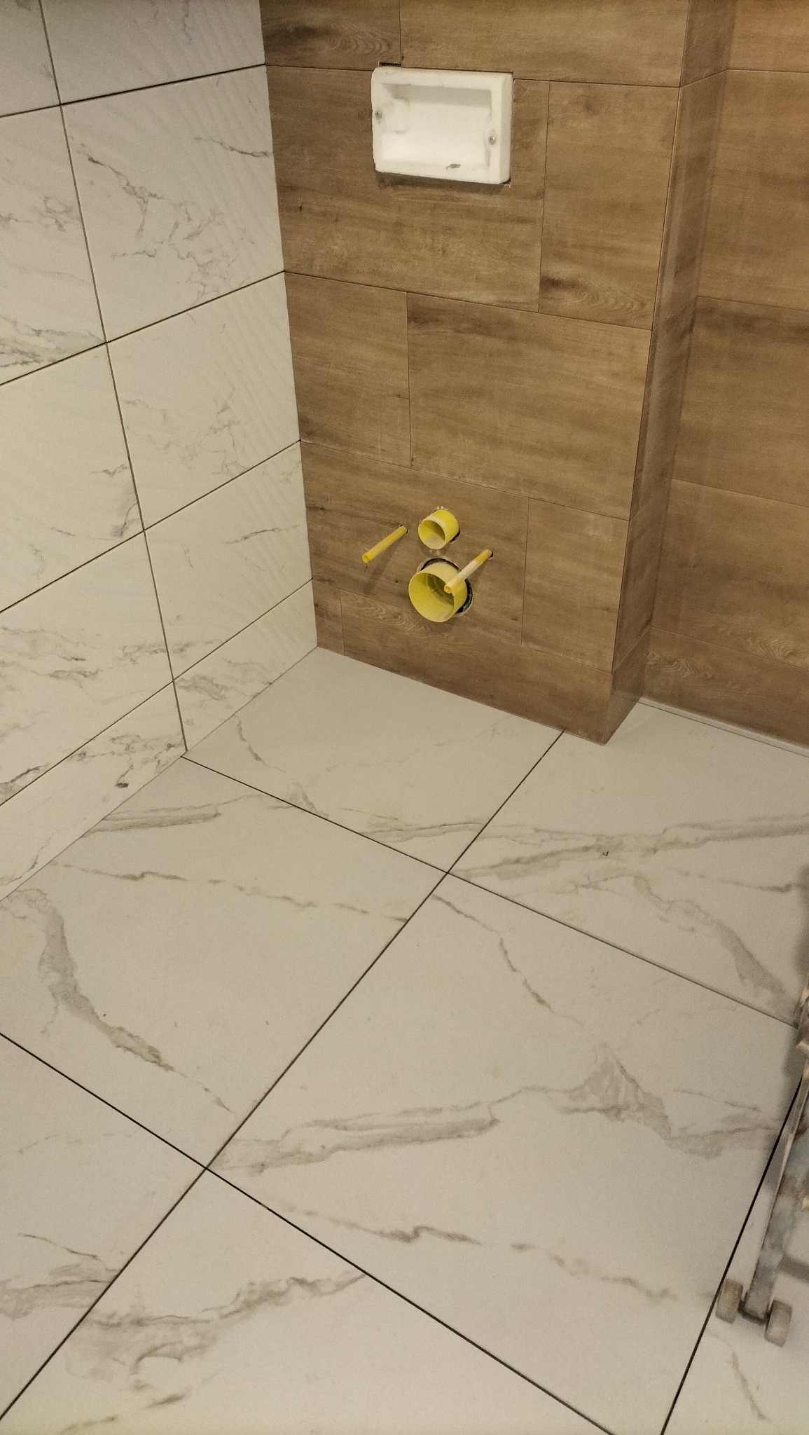 Wykończona łazienka z płytkami imitującymi marmur na podłodze i drewno na ścianach. Widoczne żółte rury i biała wnęka na ścianie. Ukończony etap prac.