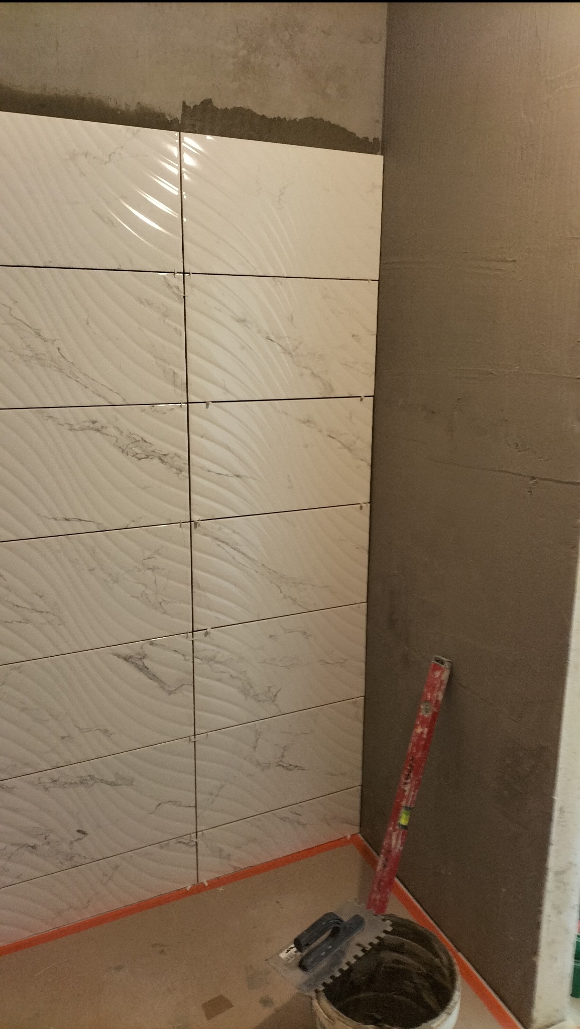 Układanie białych, wzorzystych płytek w rogu pomieszczenia. Widoczny poziomica, wiadro z zaprawą i szpachelka. Ściana przygotowana pod glazurę.