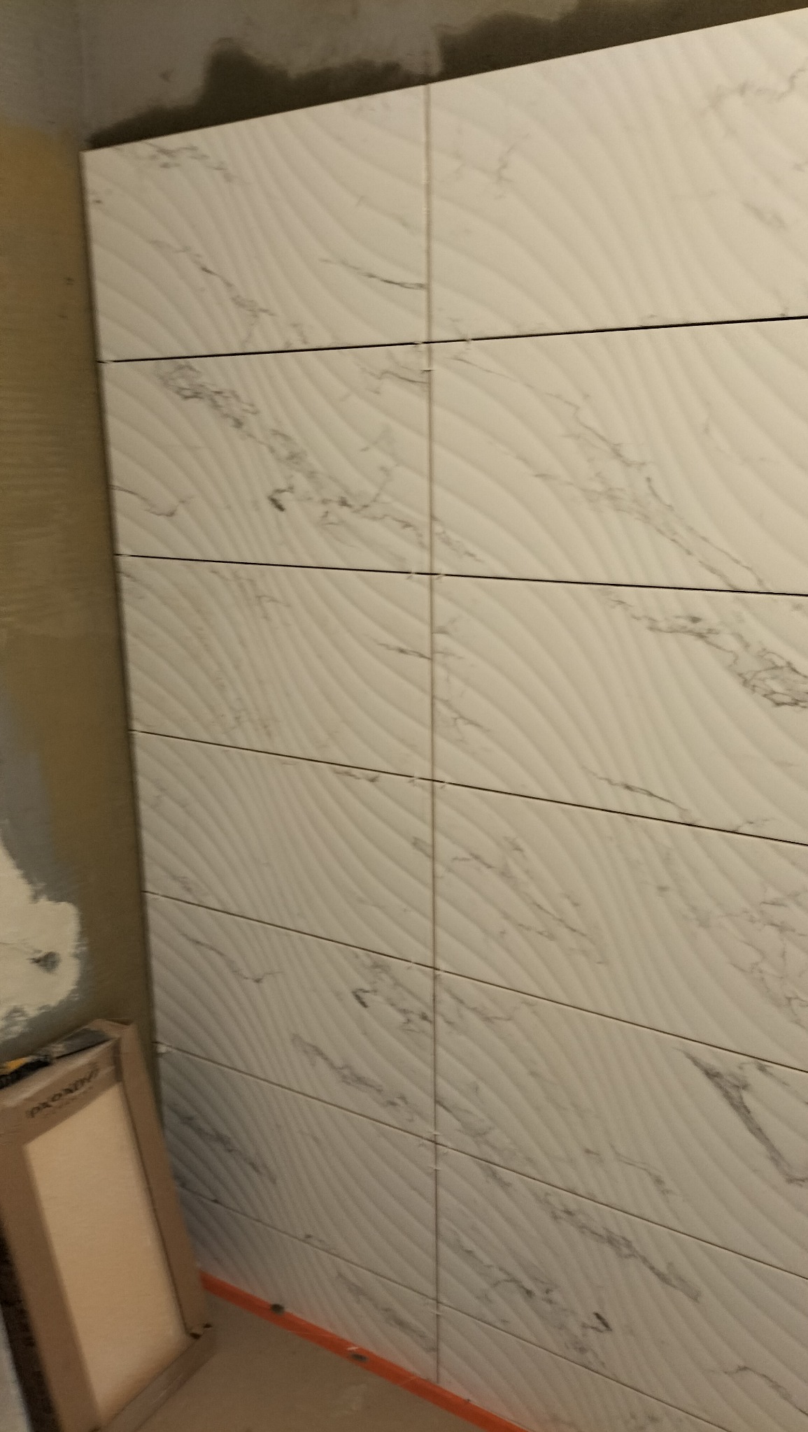 Ściana wyłożona białymi, podłużnymi płytkami z delikatnym wzorem fal i marmurkowym żyłkowaniem. Widoczne fugi i fragmenty niezakończonych prac wykończeniowych.