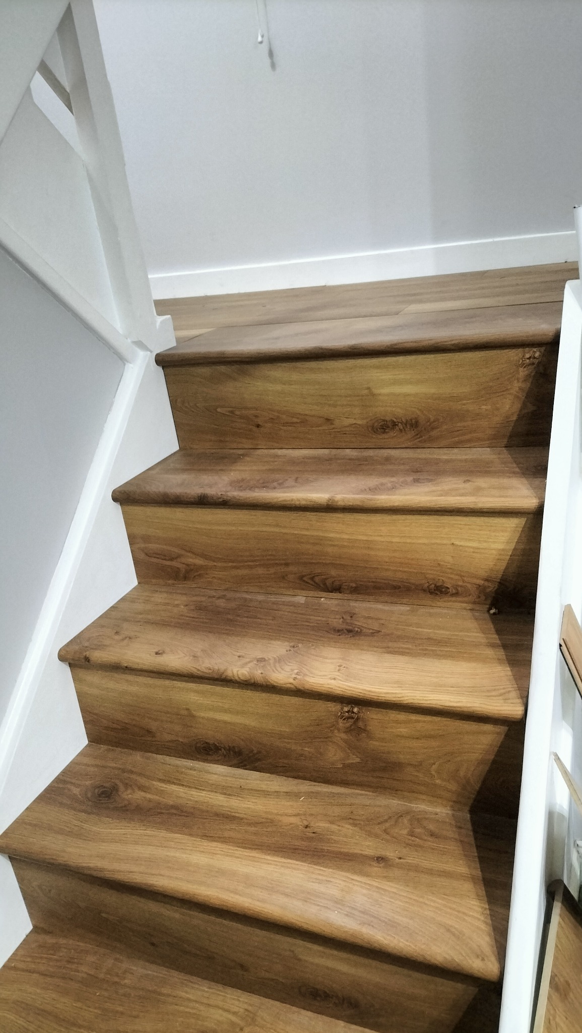 Schody wykończone panelami drewnopodobnymi, białe ściany i balustrada. Widok z dołu, ukazujący detale wykończenia i strukturę paneli.