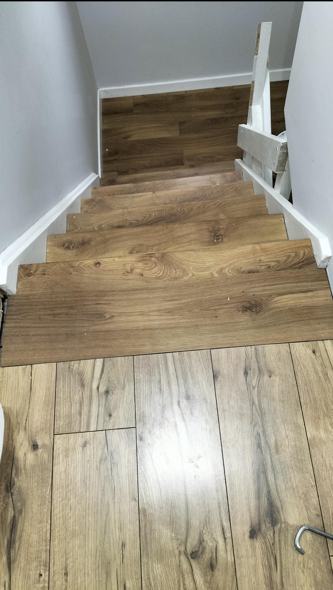 Wykończone schody z paneli drewnopodobnych, widok z góry. Jasne panele na podłodze, białe ściany i balustrada. Detal: hak narzędziowy w prawym dolnym rogu.