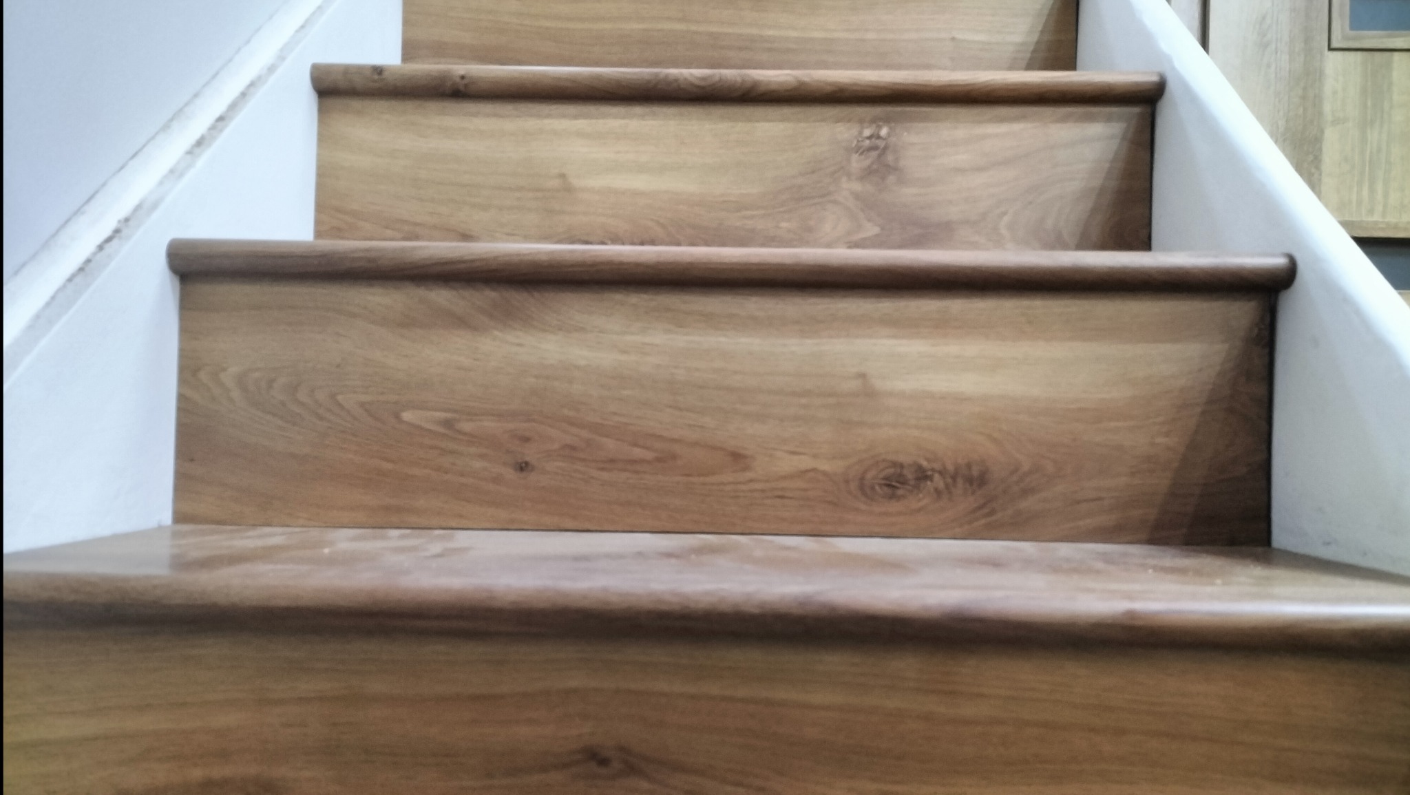 Drewniane schody z zaokrąglonymi krawędziami, widoczne z dołu, z białymi ścianami po bokach. Naturalny odcień drewna, wyraźny rysunek słojów.