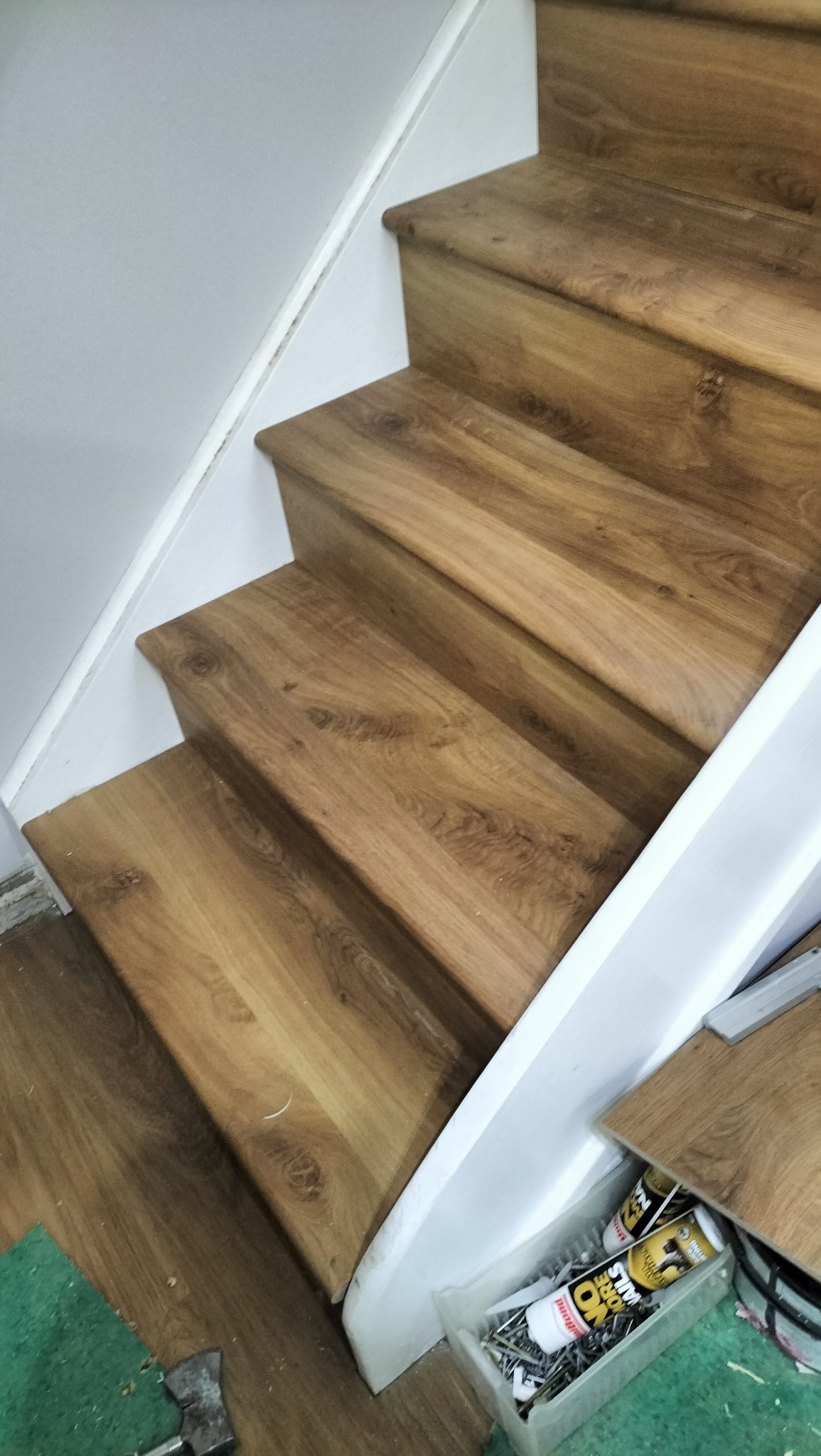 Wykończone schody z panelami drewnopodobnymi, biała barierka, widoczne narzędzia i materiały montażowe. Detal wykończenia wnętrza.