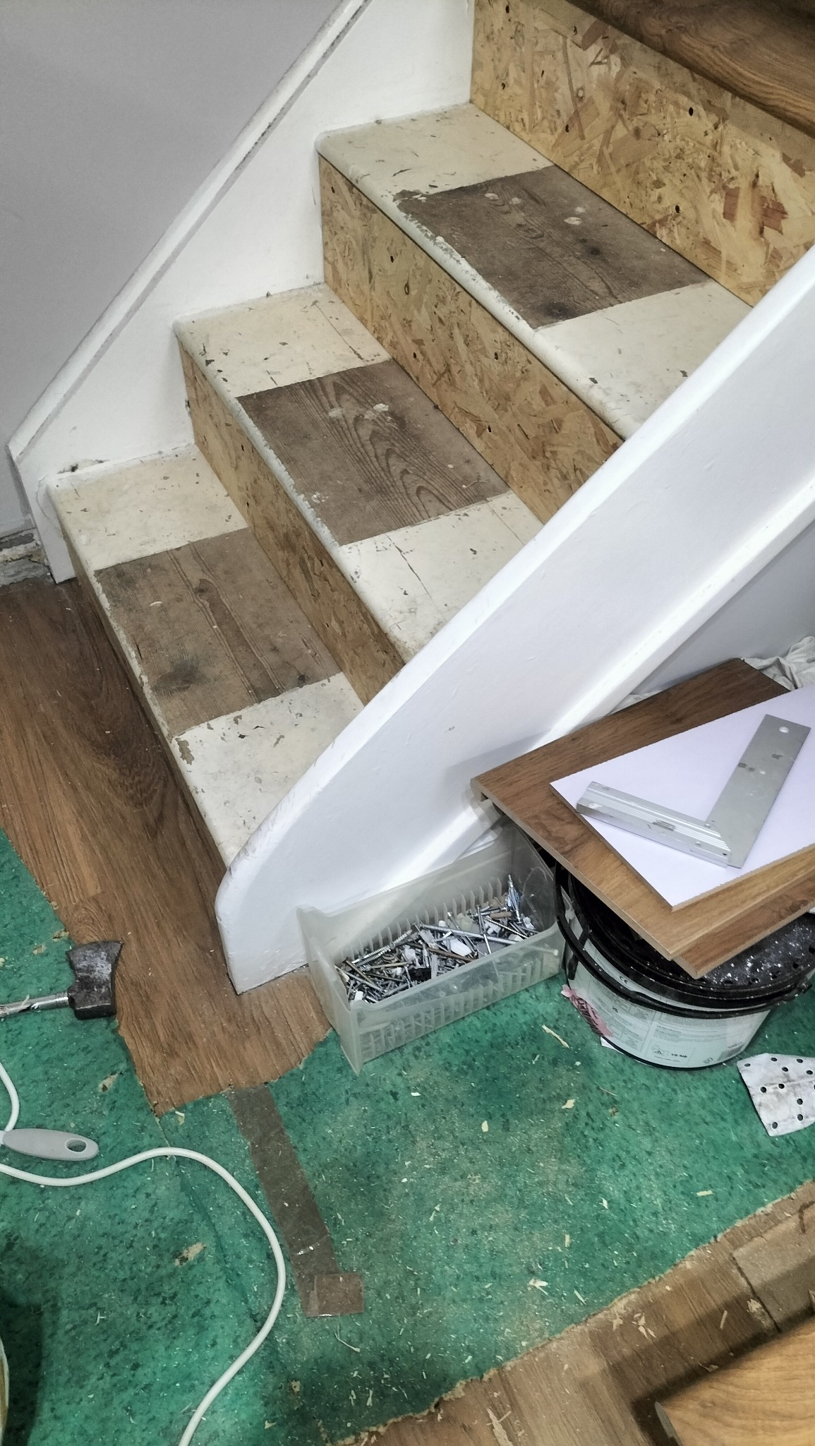 Schody w trakcie remontu: widoczne płyty OSB na stopniach, narzędzia (pojemnik z gwoździami, kątownik) i resztki materiałów budowlanych na podłodze. Etap przygotowania do wykończenia.