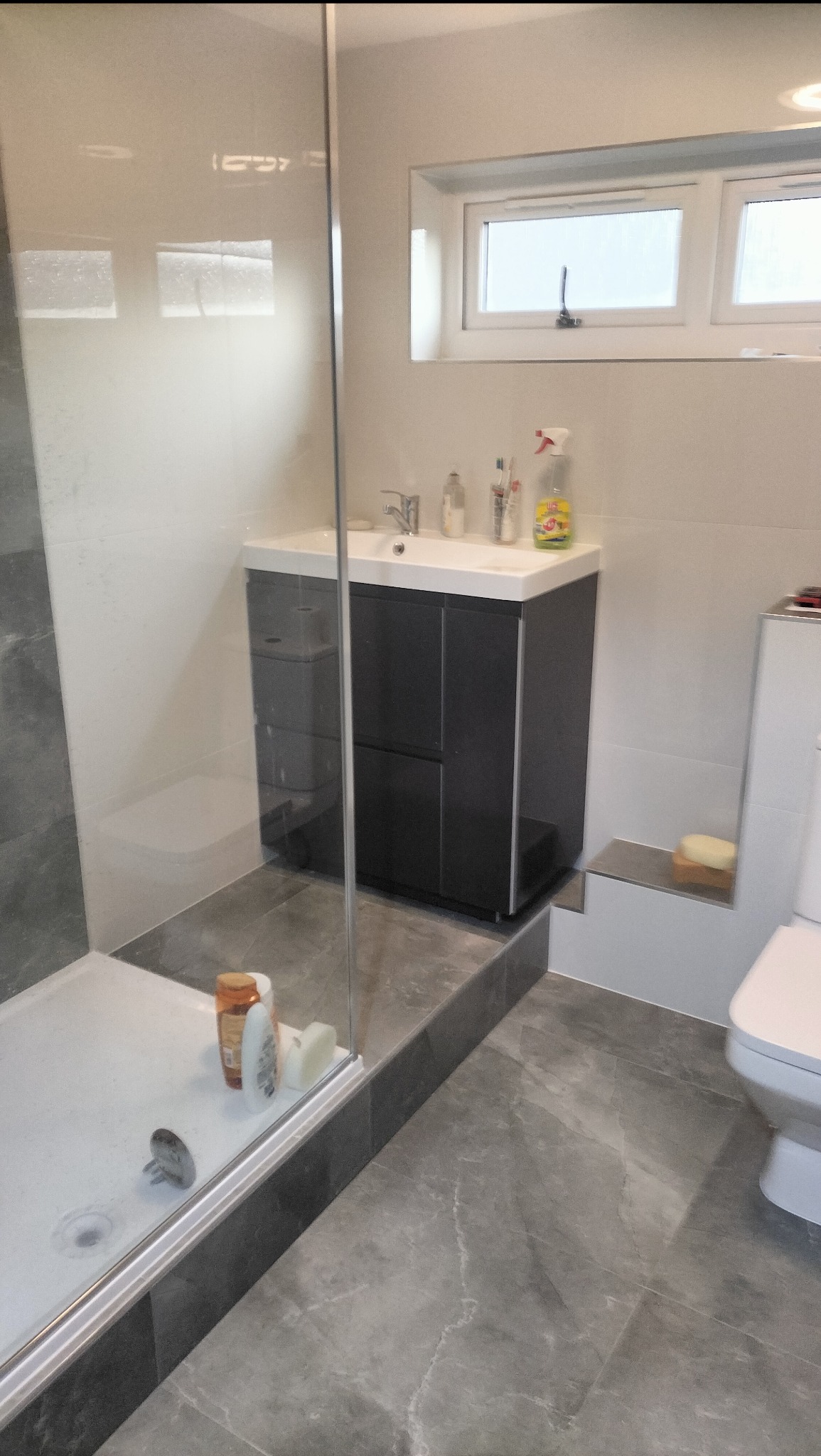 Wykończona łazienka z kabiną prysznicową, umywalką z szafką w kolorze grafitowym i szarą marmurową podłogą. Widoczne okno i akcesoria łazienkowe.