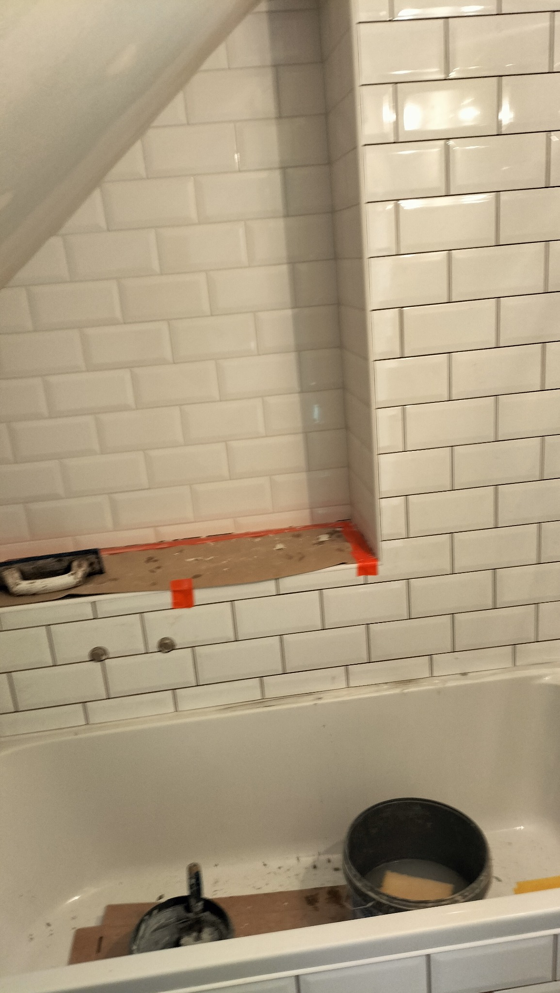 Białe, błyszczące płytki typu metro ułożone na ścianie w łazience, widoczna wanna i narzędzia glazurnicze. Półka wnękowa w trakcie wykańczania.