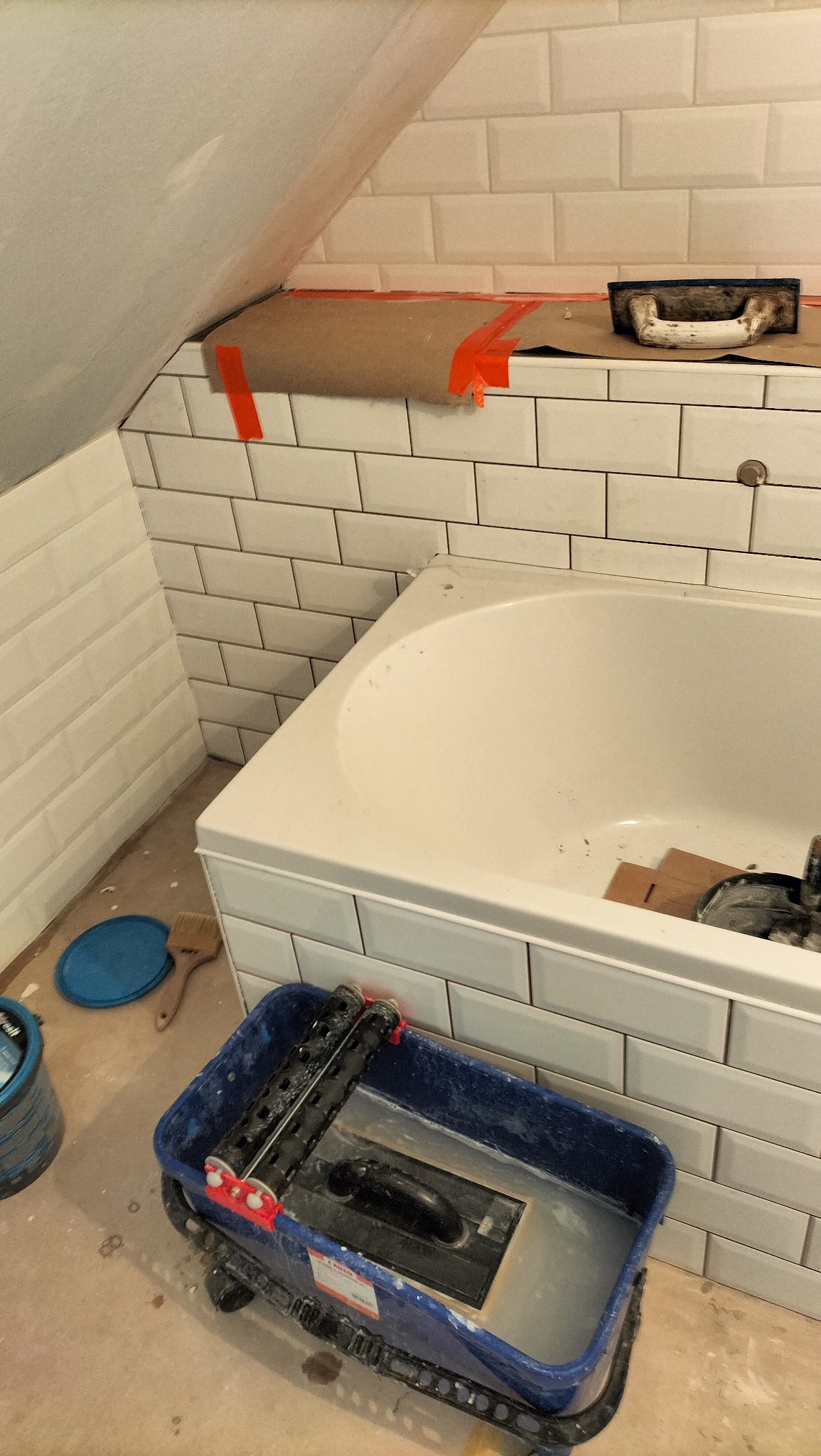 Białe, prostokątne płytki ułożone na ścianie w łazience, widoczna wanna, narzędzia glazurnicze i zabezpieczenia z kartonu i taśmy malarskiej.