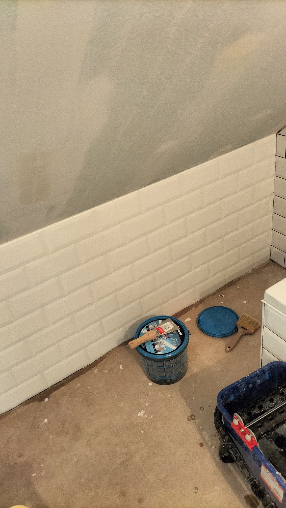 Ściana wyłożona białymi, prostokątnymi płytkami w stylu metro, z widocznym wiadrem z farbą, pędzlem i kuwetą malarską na podłodze w trakcie prac wykończeniowych.